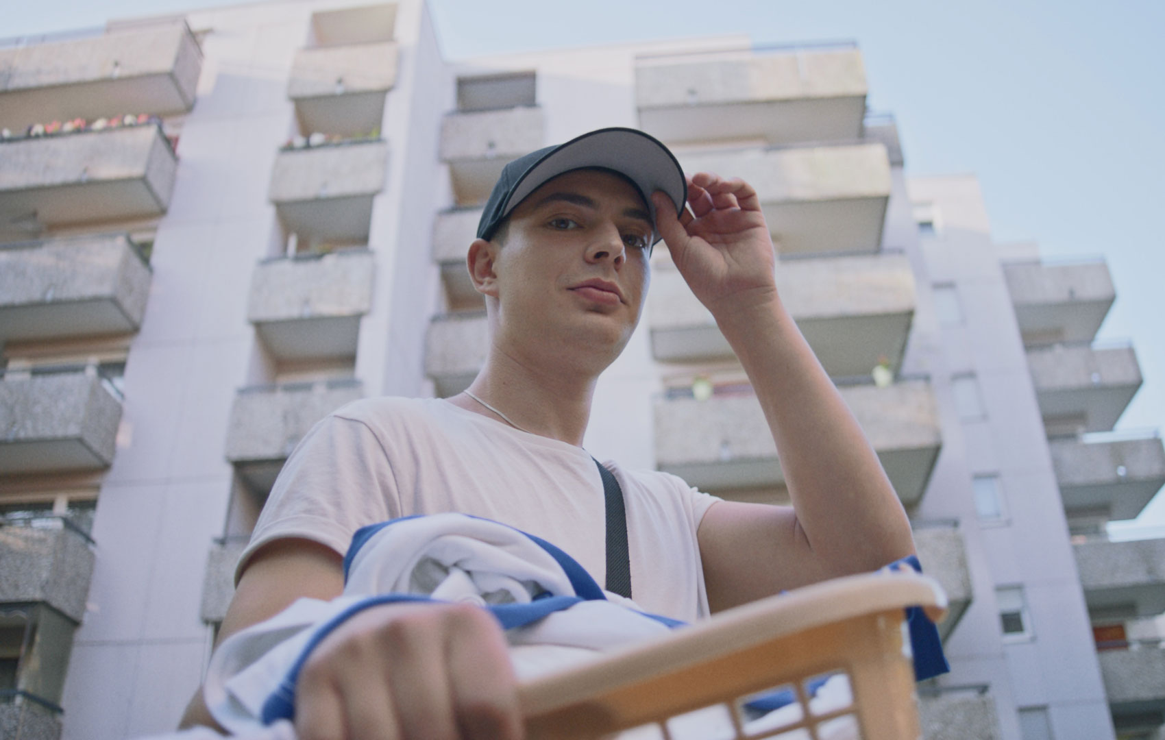 Ein Teenager vor einer Hochhaussiedlung mit Wäschekorb in der Hand. Die Kamera schaut von unten zu ihm hoch; er blickt direkt hinein.