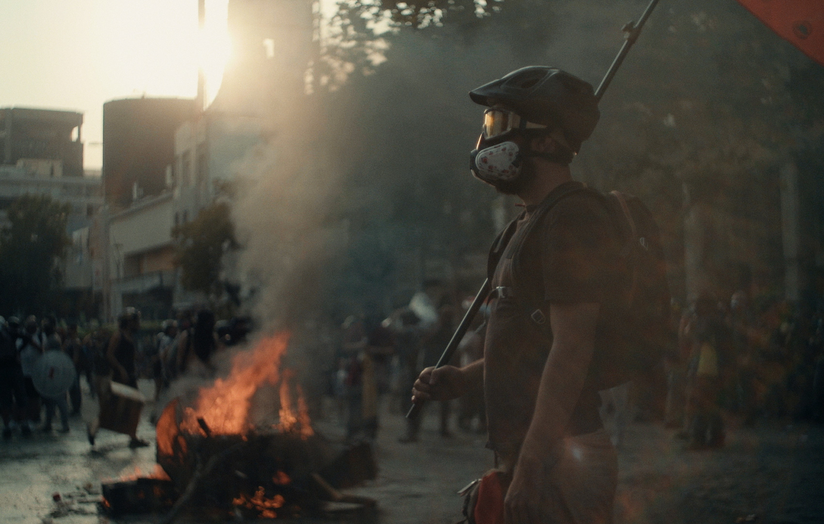 Auf einem Platz in einer Stadt brennt ein Feuer. Die Umgebung ist verraucht. Es findet eine Demonstration statt. Im Vordergrund steht ein Mann mit Helm, Schutzbrille, Atemmaske und Fahne in der Hand.