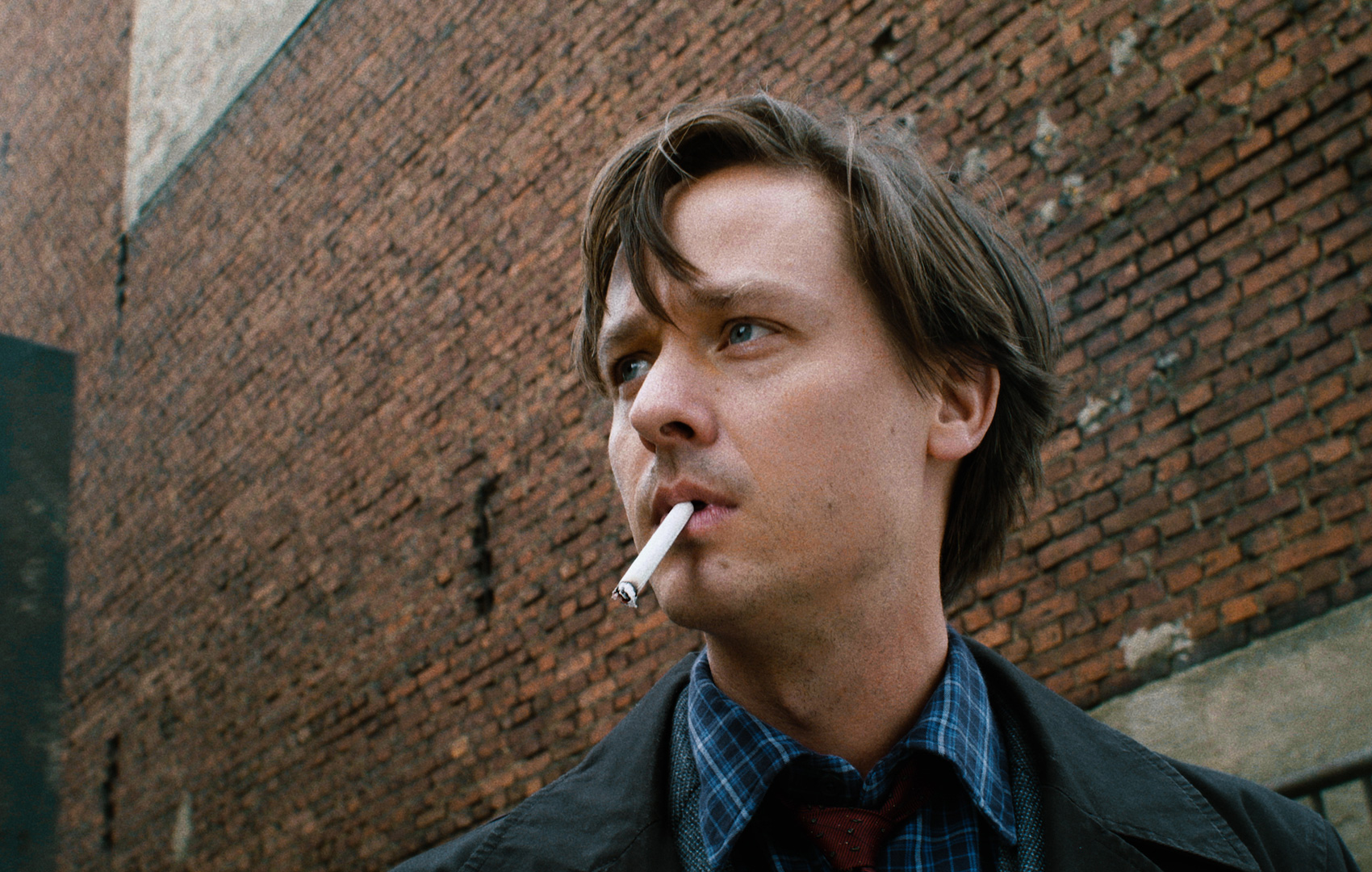 Ein von den Schultern aufwärts gezeigter Mann mit Zigarette im Mund schaut mit düsterem Blick aus dem linken Bildrand heraus.