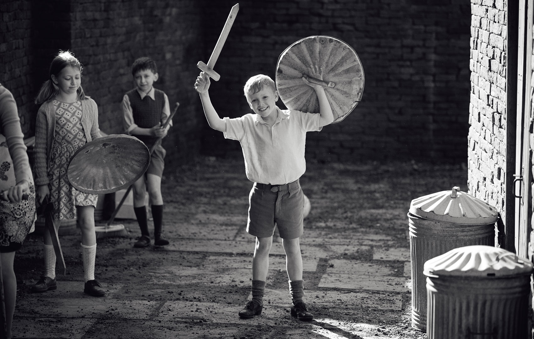 Schwarz-Weiß-Bild mit Kindern, die in einem Hinterhof mit Holzschwertern und Schutzschildern aus Mülltonnendeckeln spielen.