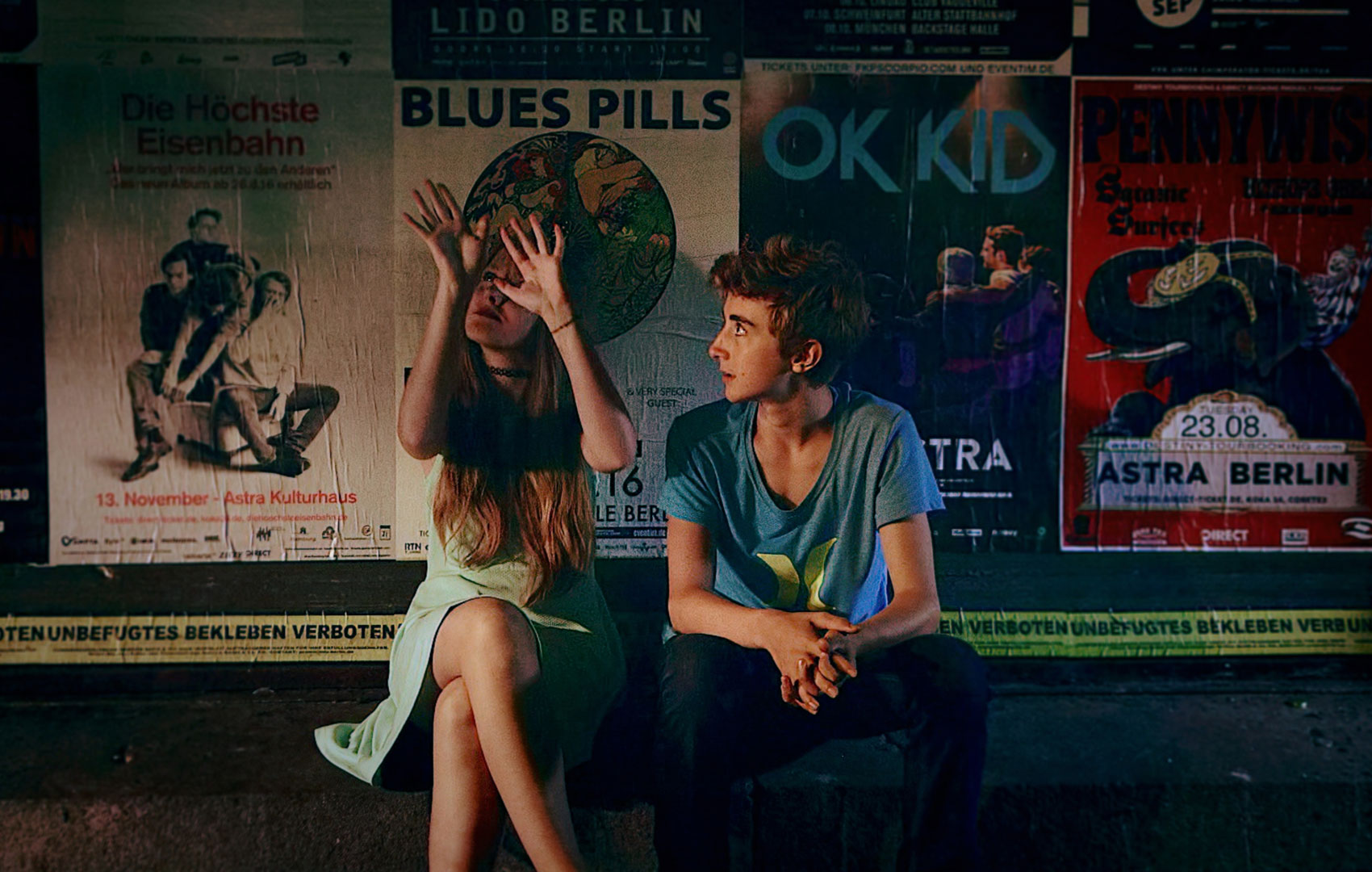 Ein Mädchen und ein Junge sitzen nebeneinader vor einer Wand mit Ankündigungspostern von Konzerten. Das Mädchen nutzt Gebärden.