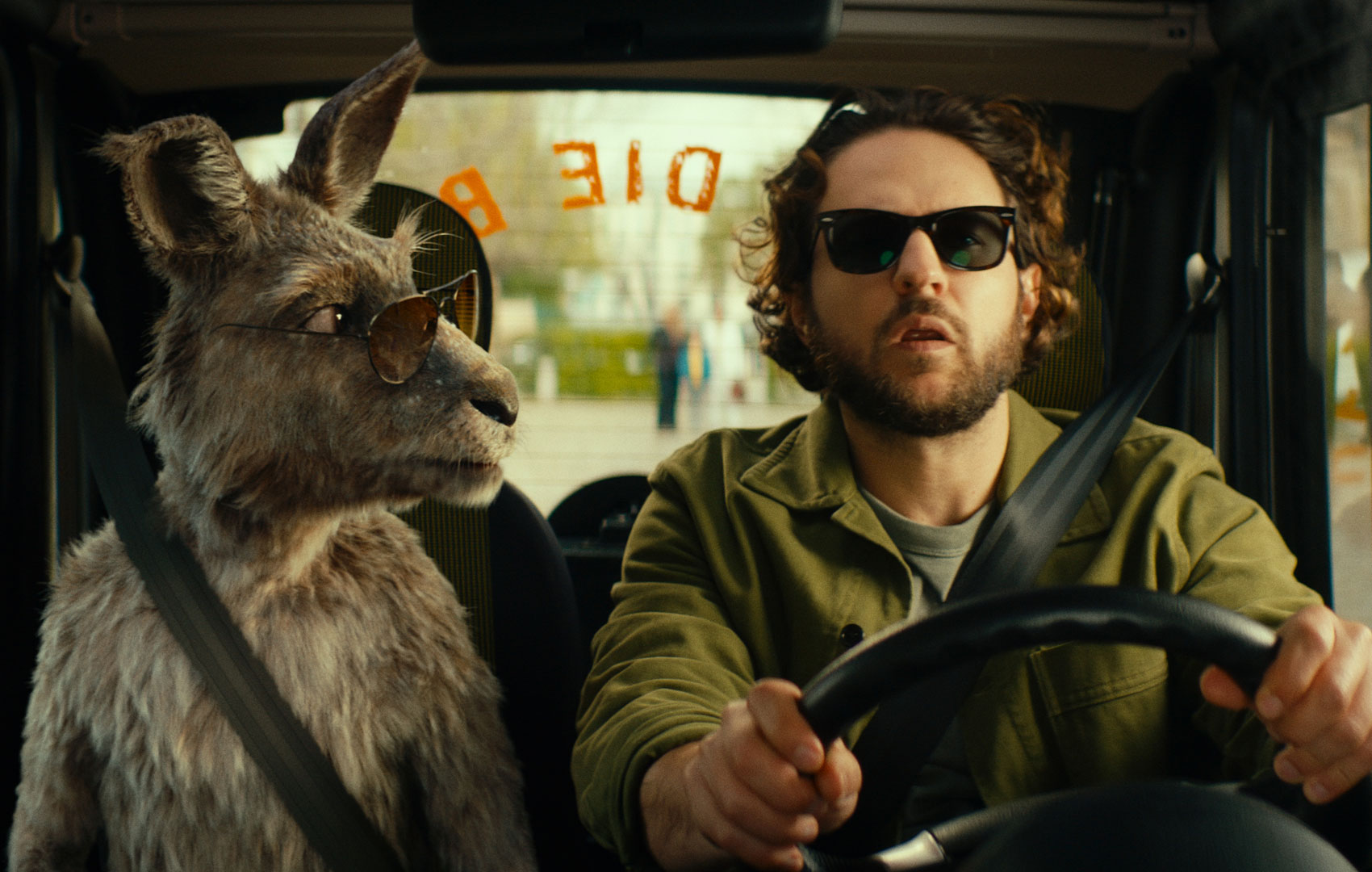 Ein Känguru sitzt links von einem männlichen Autofahrer im Auto und schaut ihn an. Beide tragen Sonnenbrillen.