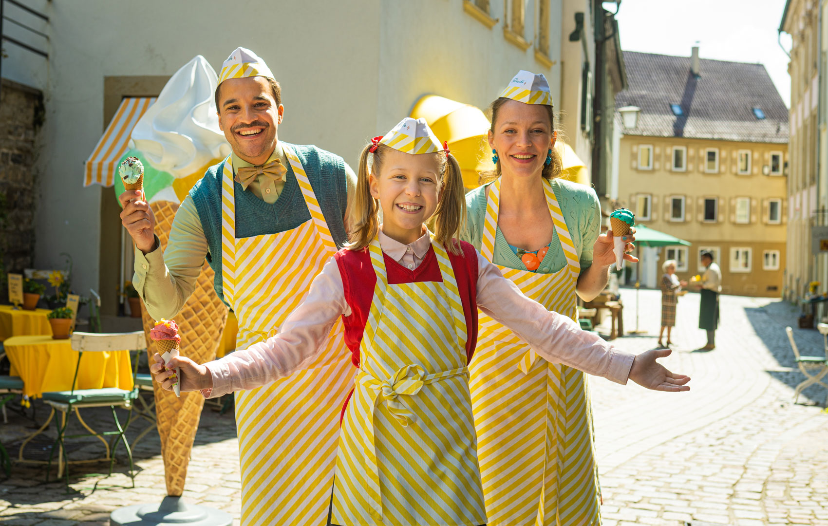 Ein Mann, eine Frau und ein Mädchen in gestreiften Schürzen halten einladend die Arme hoch. In den Händen halten sie Eiswaffeln.