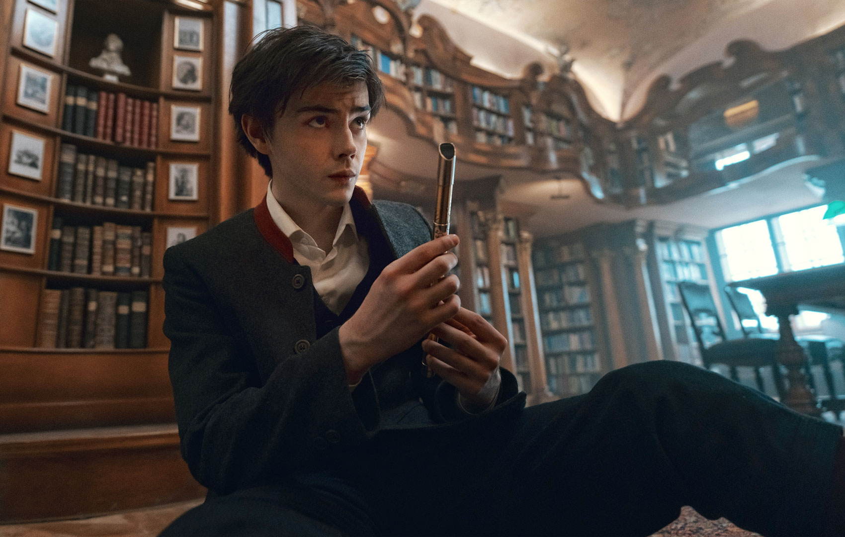 Ein Teenager in Schuluniform sitzt in einer Bibliothek auf dem Boden. Er blickt nach oben. In der Hand hat er eine kleine goldene Flöte.