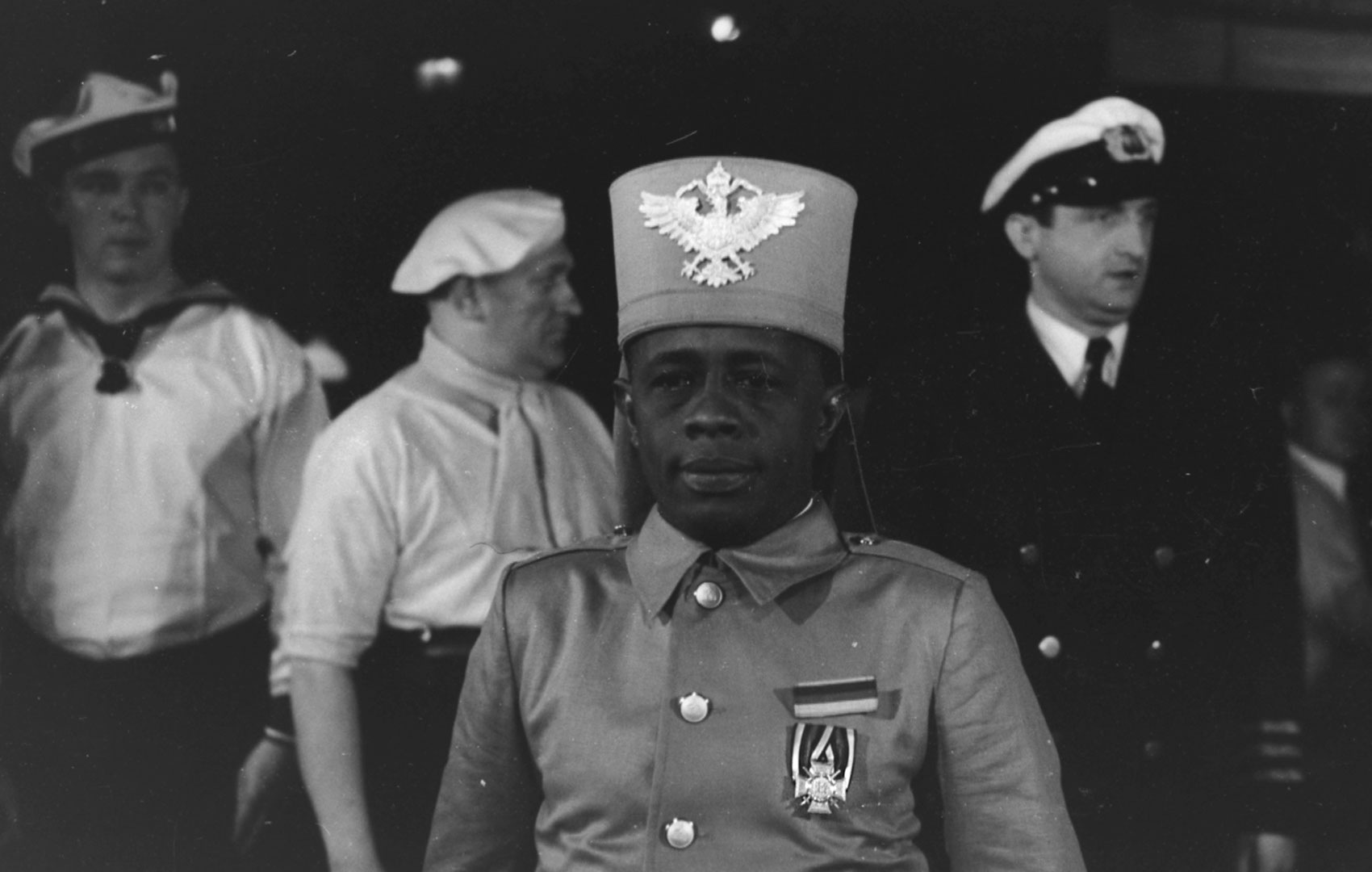Schwarz-Weiß-Bild: Im Vordergrund sieht man einen Schwarzen Mann in Kriegsuniform, im Hintergrund zwei Weiße Matrosen und ein Kapitän.