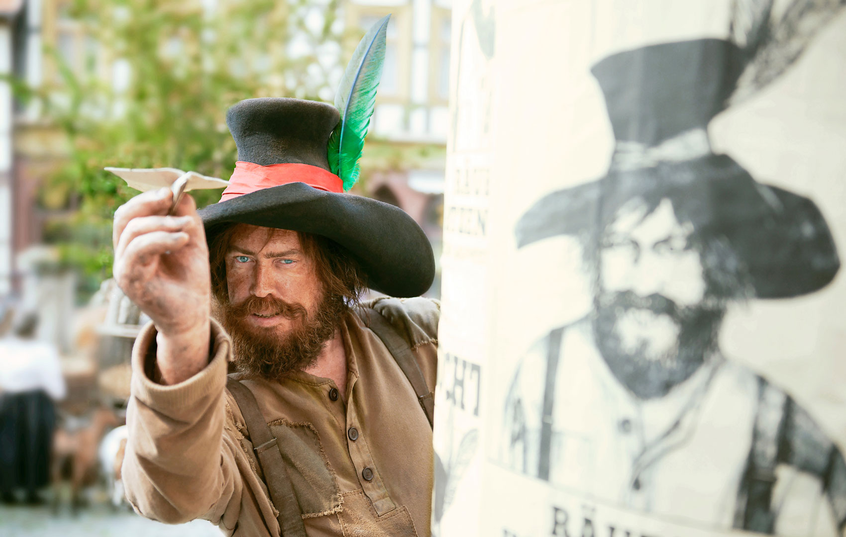 Ein Mann mit Vollbart und großem, schwarzen Hut schaut direkt in die Kamera. Er steht neben einem Plakat, auf dem er mit Phantombild von der Polizei gesucht wird.