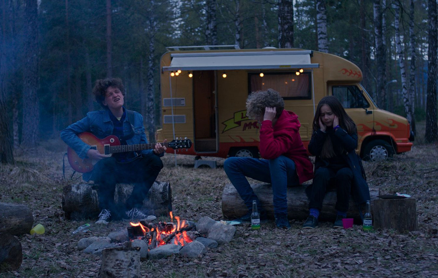 Drei Freund:innen unterschiedlichen Alters sitzen an einem Lagerfeuer im Wald. Zwei von ihnen halten sich die Ohren zu, während einer Gitarre spielt und singt.