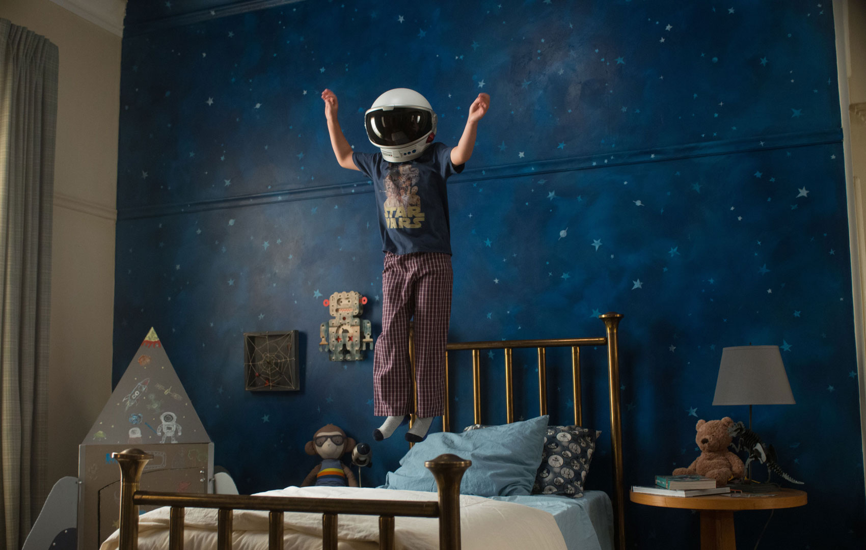 Ein Kind mit Astronautenhelm auf dem Kopf hüpft auf seinem Bett vor einer Weltraum-Sternen-Tapete.