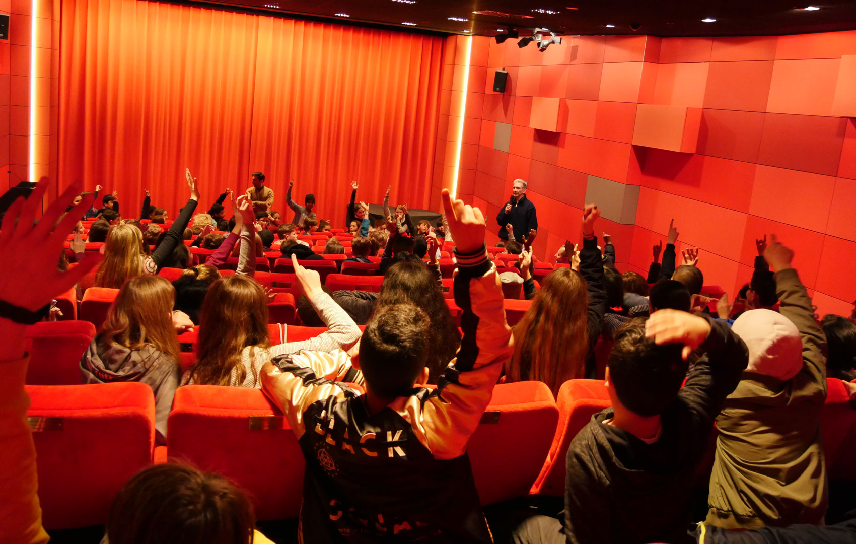 Kinosaal mit Kindern, die die Hände hochheben, um Fragen zu stellen. Vor der Leinwand stehen zwei Männer mit Mikrofonen in der Hand.