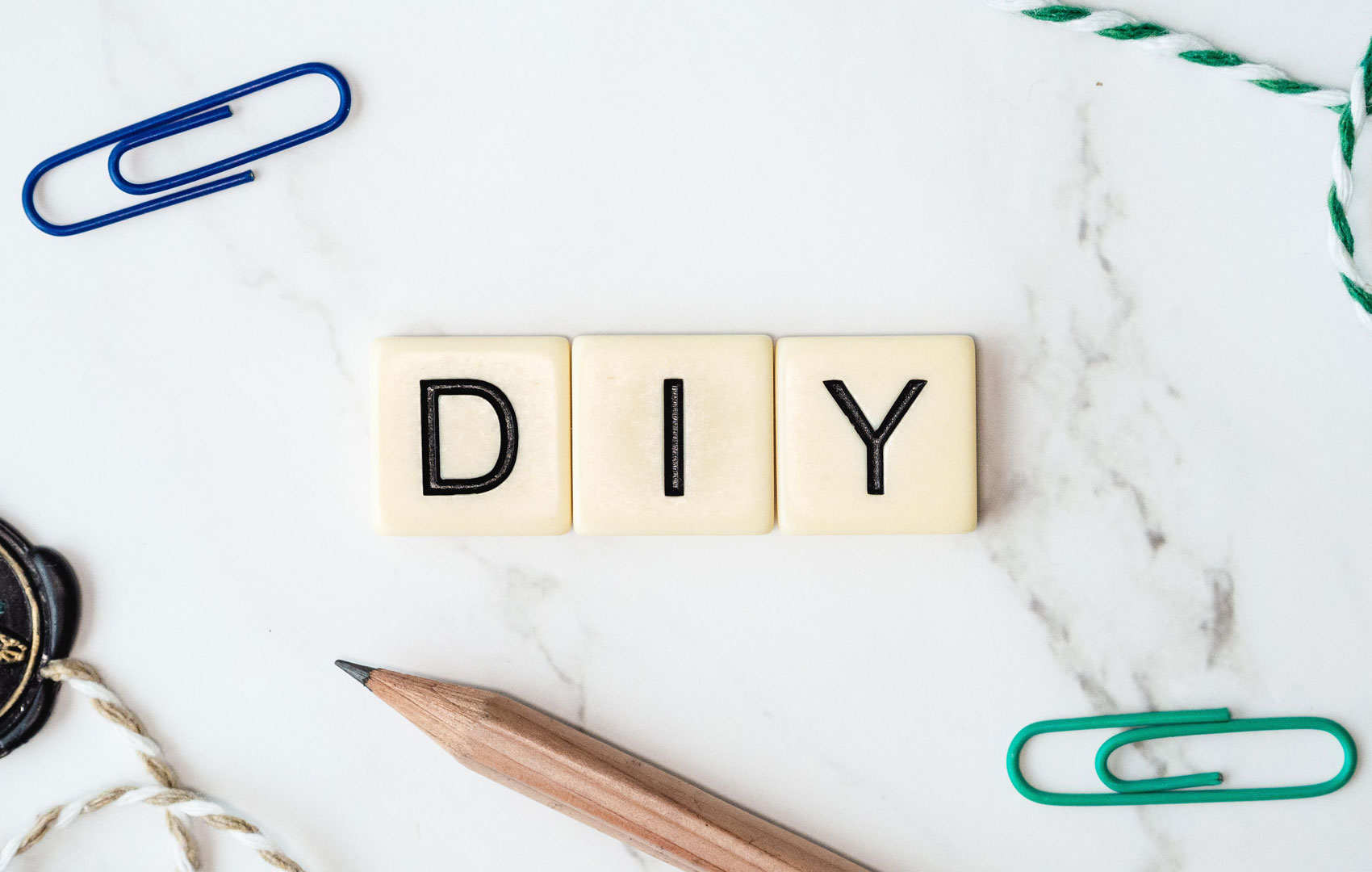 Drei Scrabble-Buchstabensteine formen das Wort D I Y. Außenherum sind ein Bleistift, Büroklammern und Fäden zu sehen.