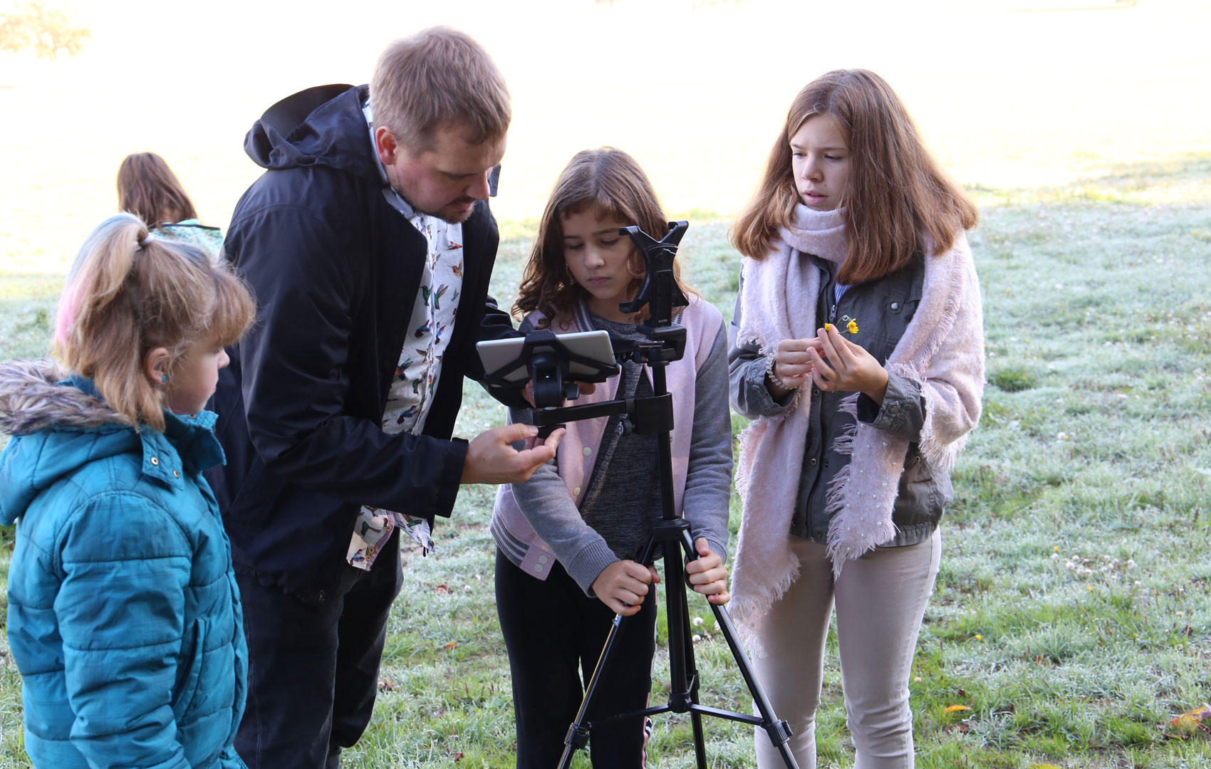 Ein Mann steht mit jungen Mädchen auf einer Wiese und und sie blicken gemeinsam auf ein Tablet, das in ein Stativ eingefasst ist.