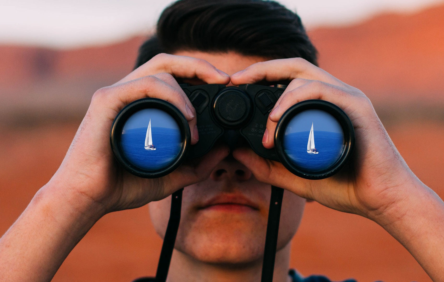 Ein Mann schaut durch ein Fernglas. In den Gläsern sieht man rechts wie links jeweils blaues Meer mit einem weißen Segelboot darauf.