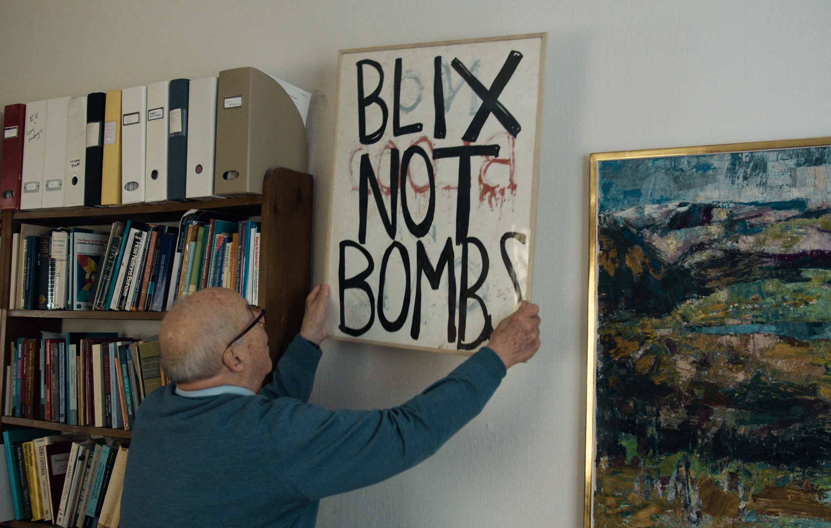 Älterer Mann hängt zwischen ein Bücherreal und ein Gemälde ein Schild an die Wand auf dem in Großbustaben BLIX NOT BOMBS steht.