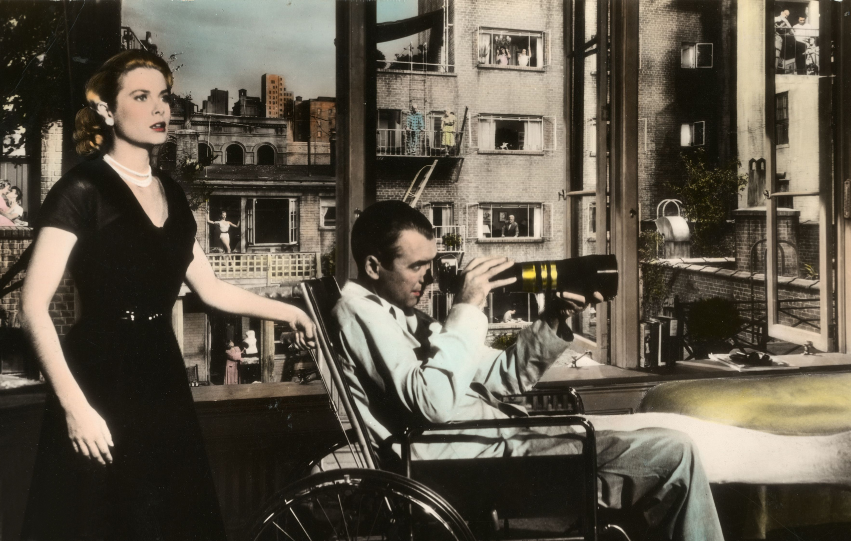 Frau mit erschrockenem Blick links im Bild hat eine Hand am Schiebegriff eines Rollstuhls. Darin sitzt ein Mann mit einem Gipsbein. In der Hand hält er eine Fotoapparat mit Teleobjektiv. Im Hintergrund ist eine Häuserfront mit Fenstern zu sehen.