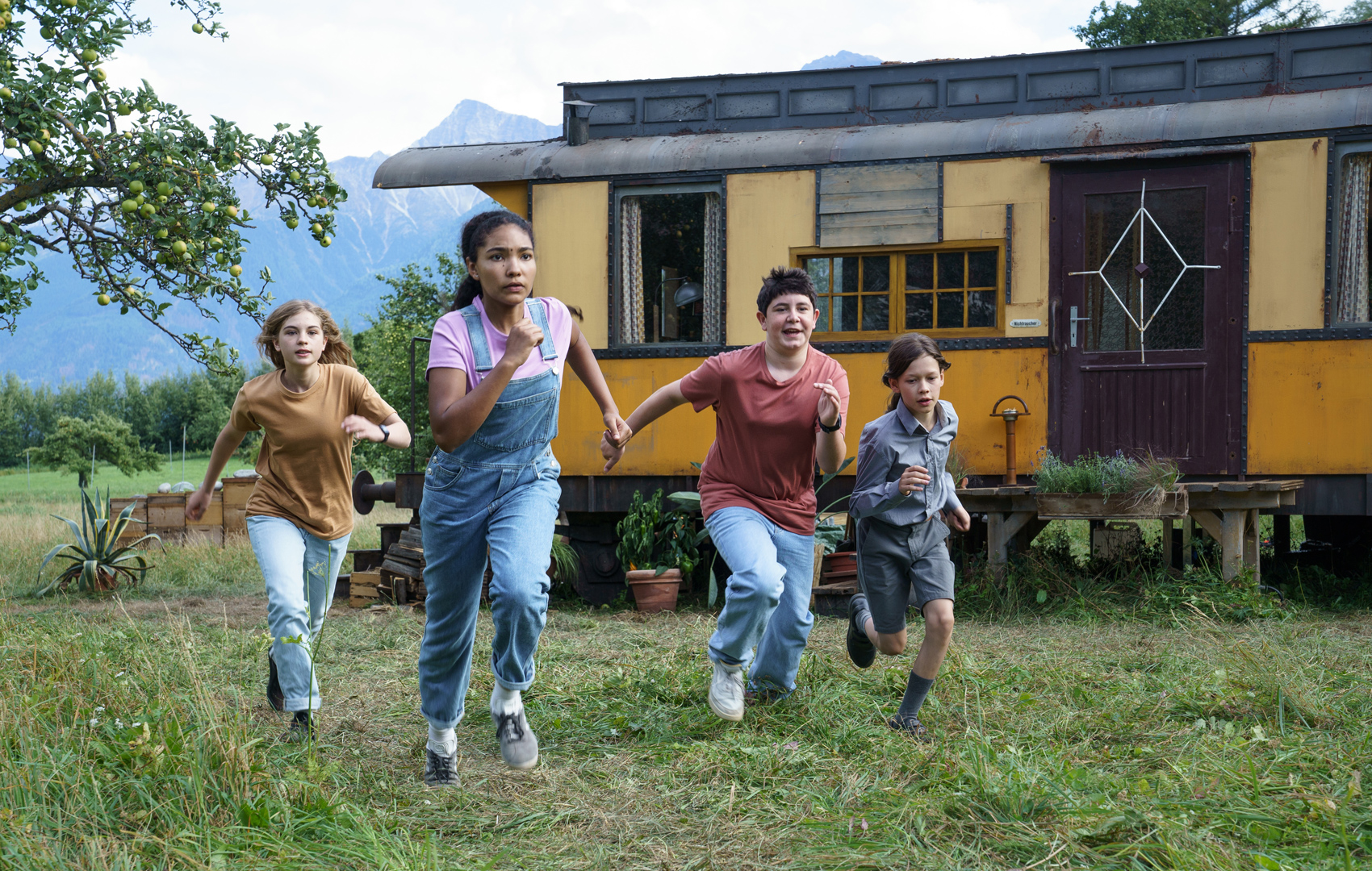 Vier Kinder auf einer Wiese rennen in Richtung des Bildvordergrunds. Im Hintergrund sieht man einen alten Zugwagon, der auf einer Wiese in einem großen Garten steht.