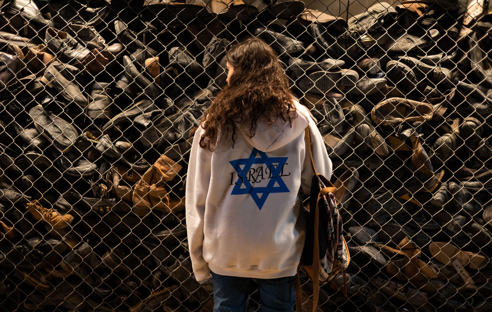 Eine junge Frau steht vor einem Maschendrahtzaun, hinter dem Schuhe gestapelt sind. Sie trägt einen weißen Pullover mit dem blauen Stern der israelischen Flagge. Das Wort ISRAEL ist in Großbuchstaben quer darüber geschrieben.