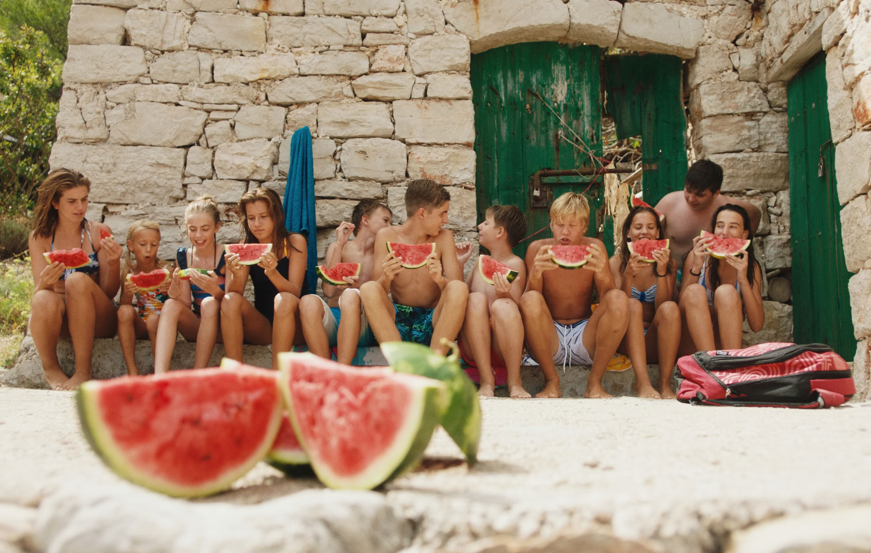 Im Hintergrund vor einem alten Steinbauernhaus sitzen elf Kinder in Badekleidung auf einer Steinstufe und essen Wassermelonenschnitze.