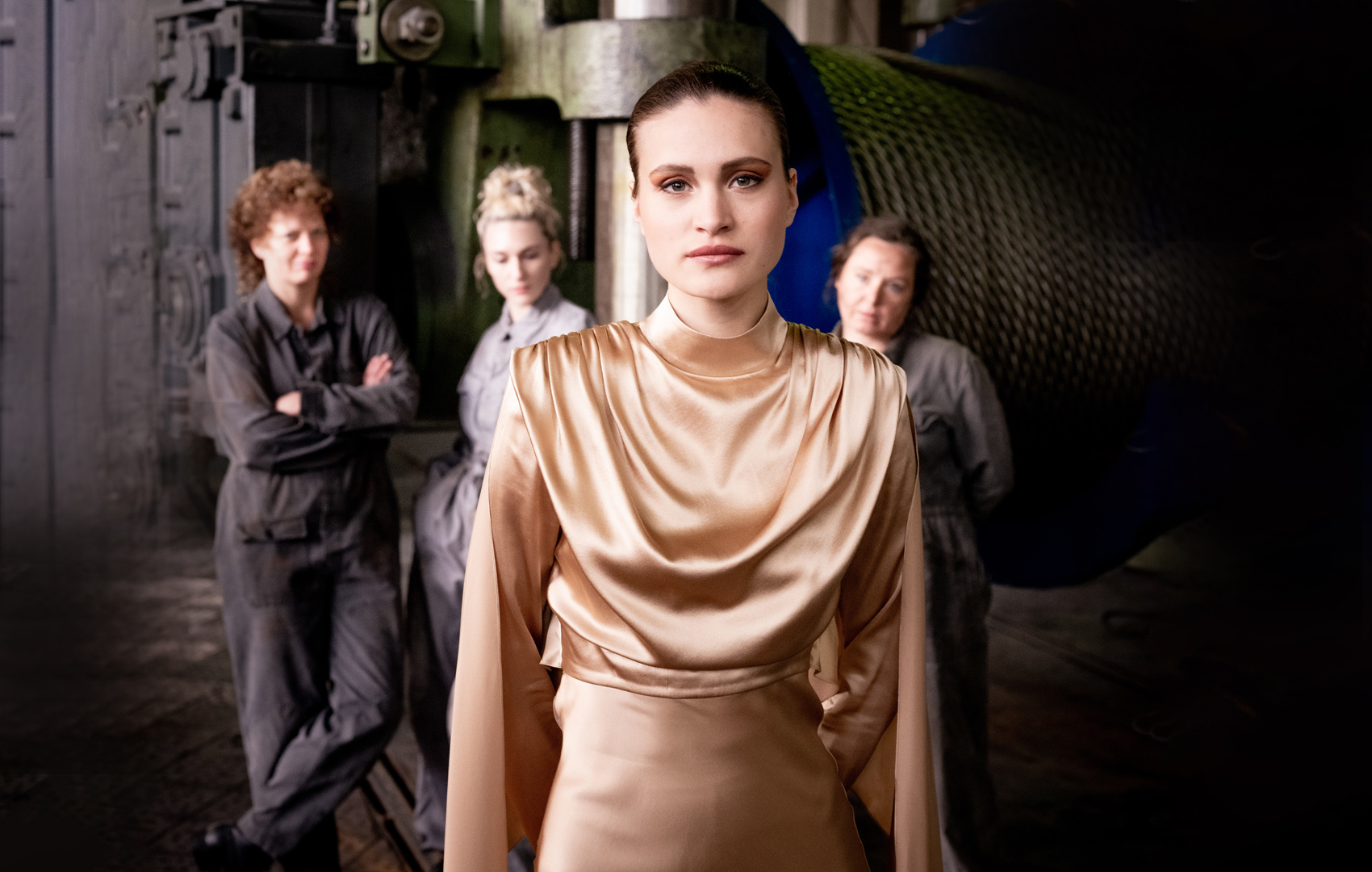 Eine junge Frau in chicen goldenen Kleid steht in einer Fabrik. Im Hintergrund sind ihre drei Kolleginnen in grauen Overalls zu sehen.