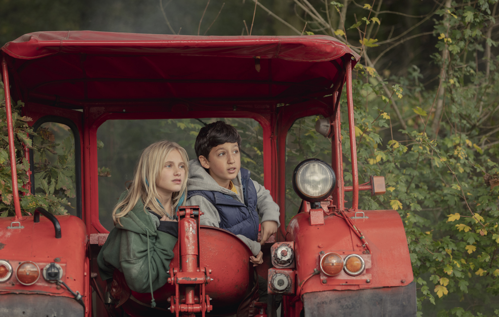 Ein Mädchen und ein Junge sitzen in einem alten roten Traktor vor einem buschigen Wald. Sie schauen aus dem Bild hinaus.