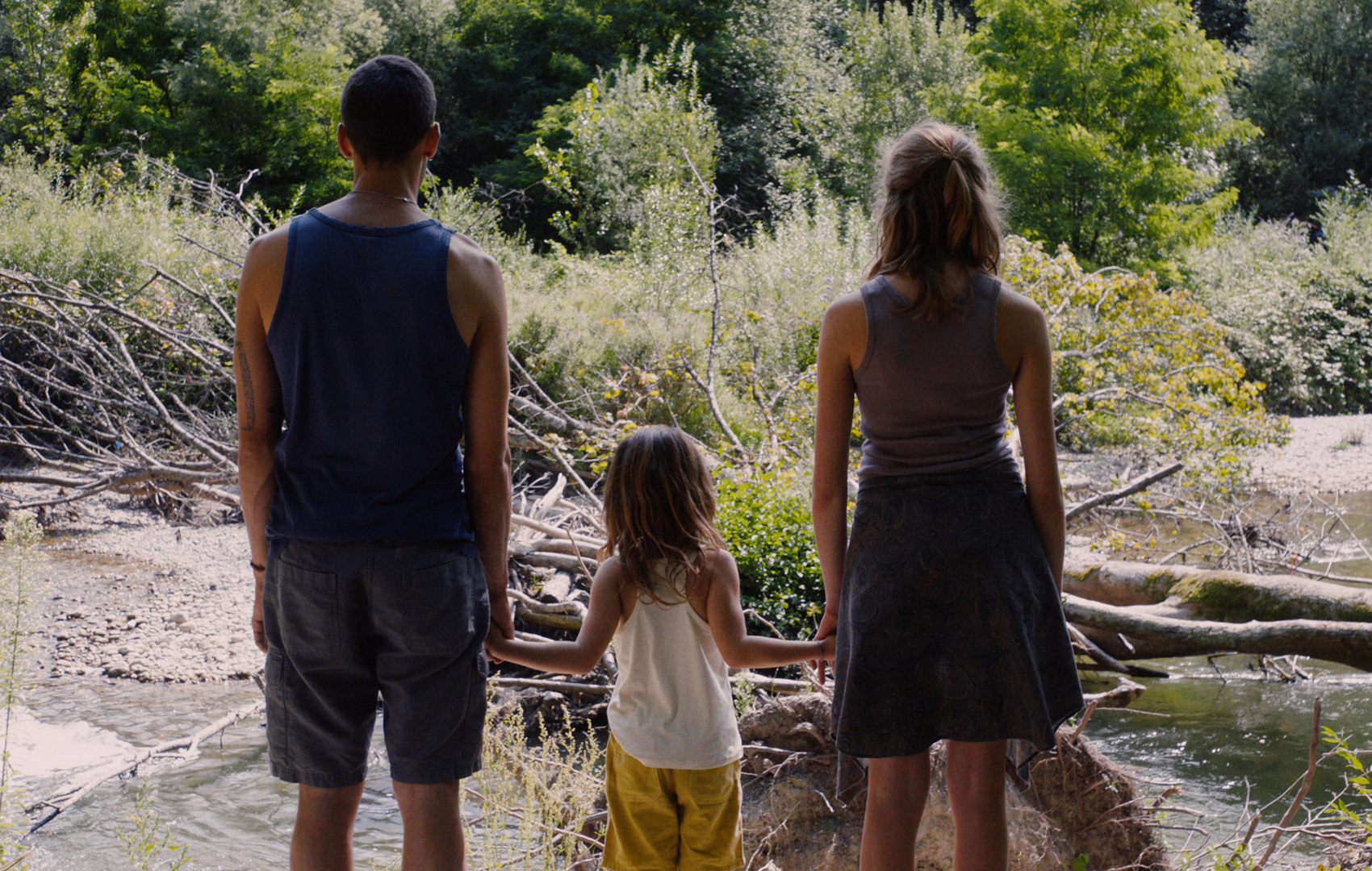 Ein Mann, ein kleines Mädchen und eine Jugendliche halten sich an den Händen und blicken auf eine Waldlandschaft mit Fluss. Sie stehen mit dem Rücken zur Kamera.