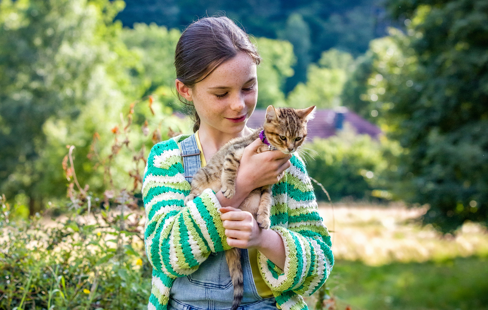 Ein Mädchen hält eine kleine Katze auf dem Arm. Sie stehen in einem großen Garten oder Waldgebiet.