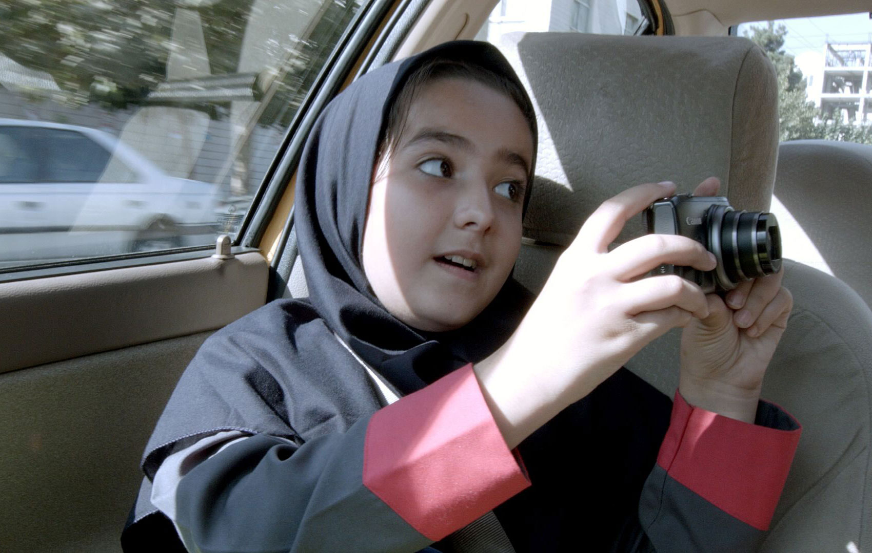 Ein Mädchen mit Kopftuch sitzt auf dem Beifahrersitz eines Autos. Sie blickt mit einer Digitalkamera in der Hand zum Autofahrer außerhalb des Bildes.