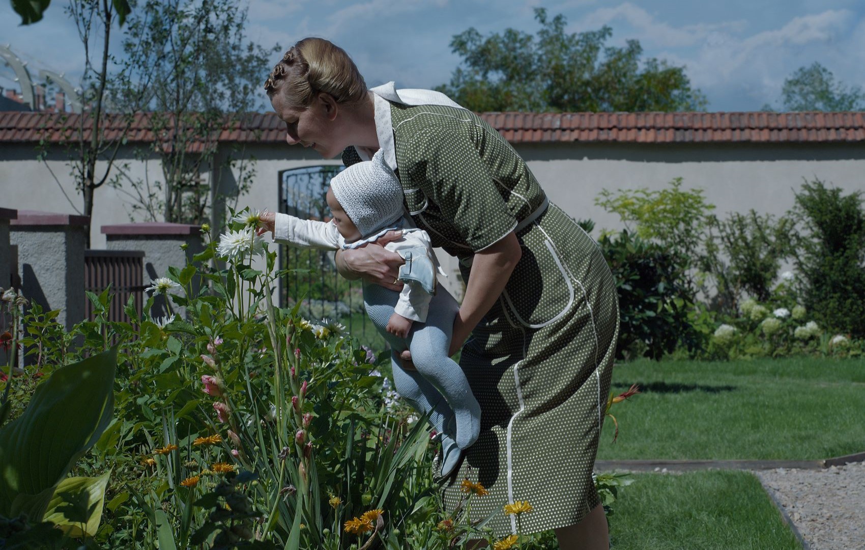 Im Garten hält eine Frau ein Baby im Arm. Das Baby greift nach einer offenen Blume. Die Kleidung der beiden ist aus den 1940er Jahren.