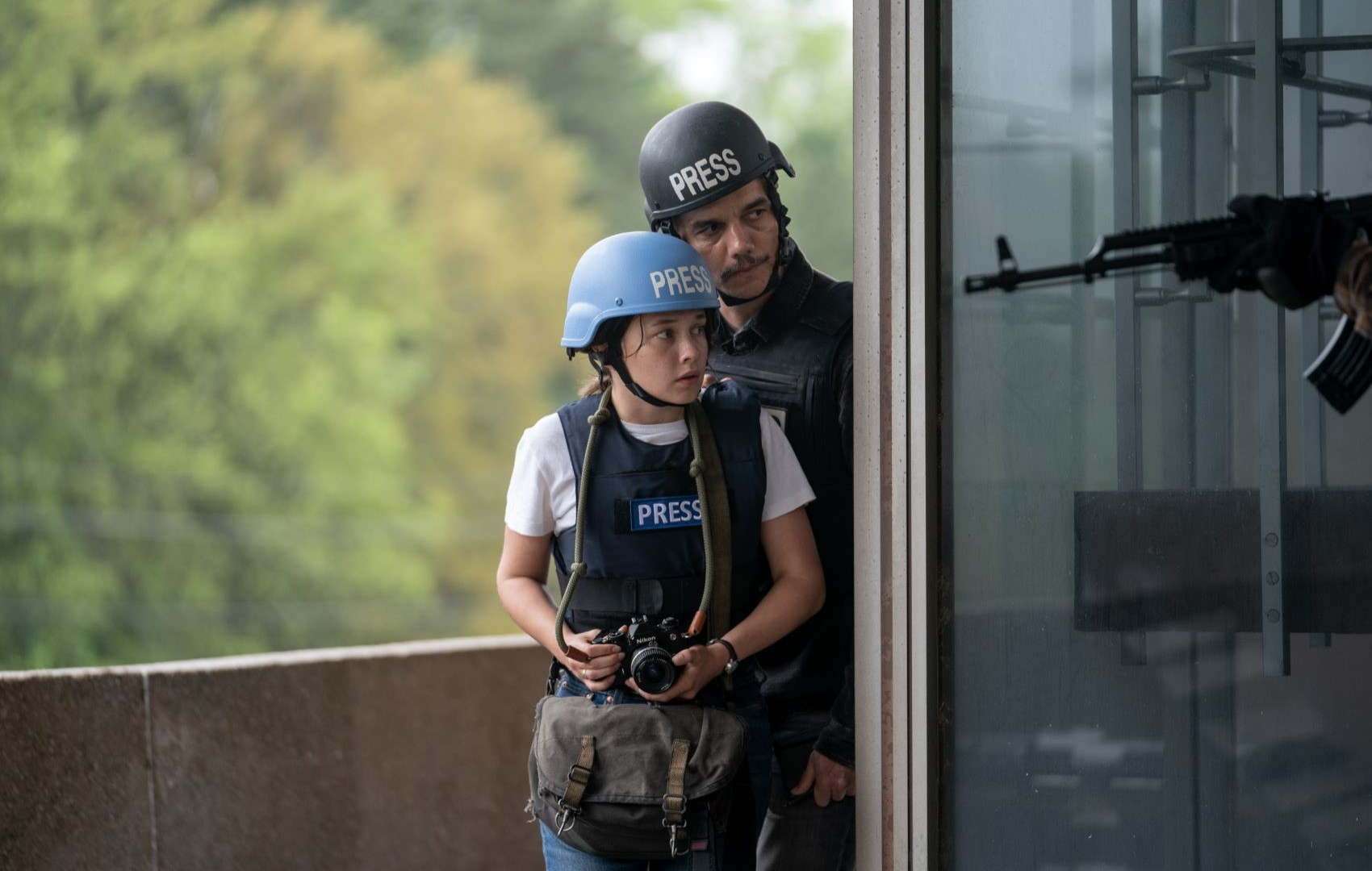 Ein Mann und eine junge Frau sind zu sehen. Sie hat eine Fotokamera dabei und beide tragen einen Schutzhelm und kugelsichere Westen.