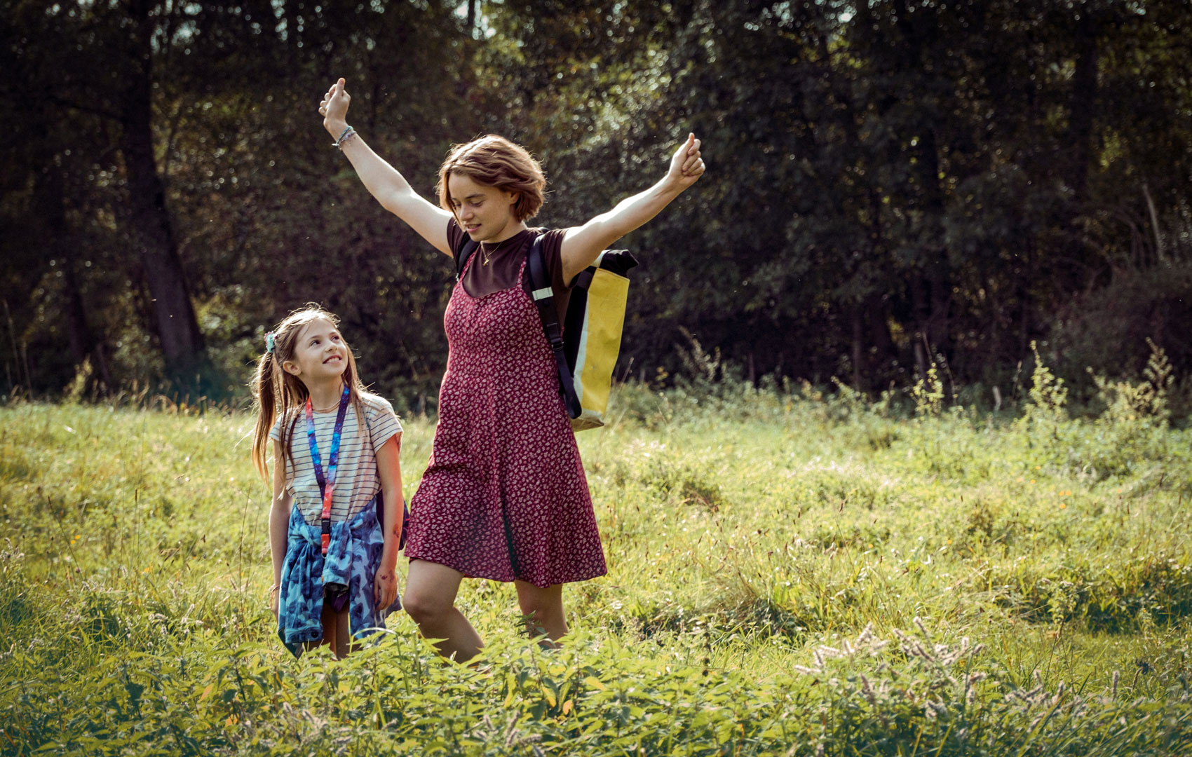 Zwei Mädchen laufen über eine Sommerwiese. Das jüngere schaut lächelnd zum älteren Mädchen auf, das gerade seine Arme in die Luft streckt, als hätte es Flügel.
