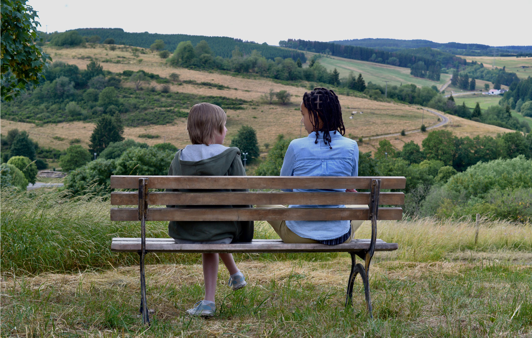 Ein Junge und ein Mädchen sitzen mit dem Rücken zum Publikum auf einer Bank in der Natur. Sie schauen sich an; im Hintergrund ist eine Berg- und Feldlandschaft zu sehen.