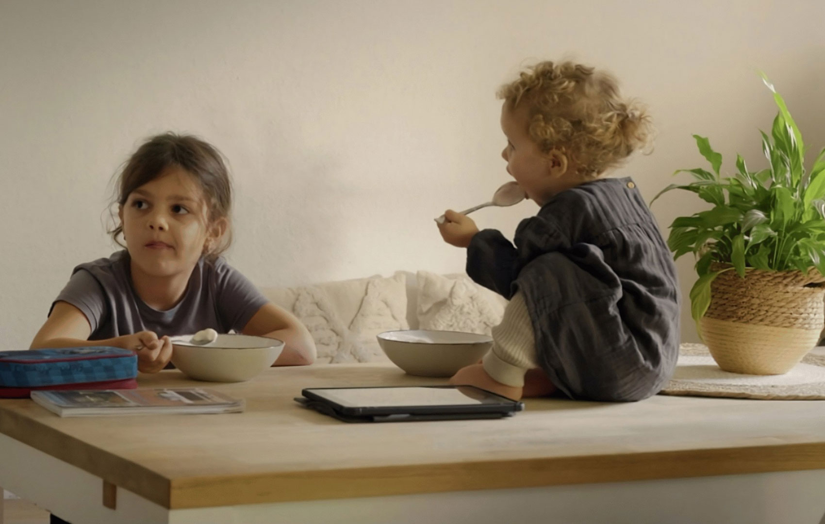 Zwei junge Kinder sitzen an einem Tisch. Eines auf einem Stuhl, das andere sitzt im halb gegenüber auf dem Tisch. Beide löffeln etwas auf Schüsseln.