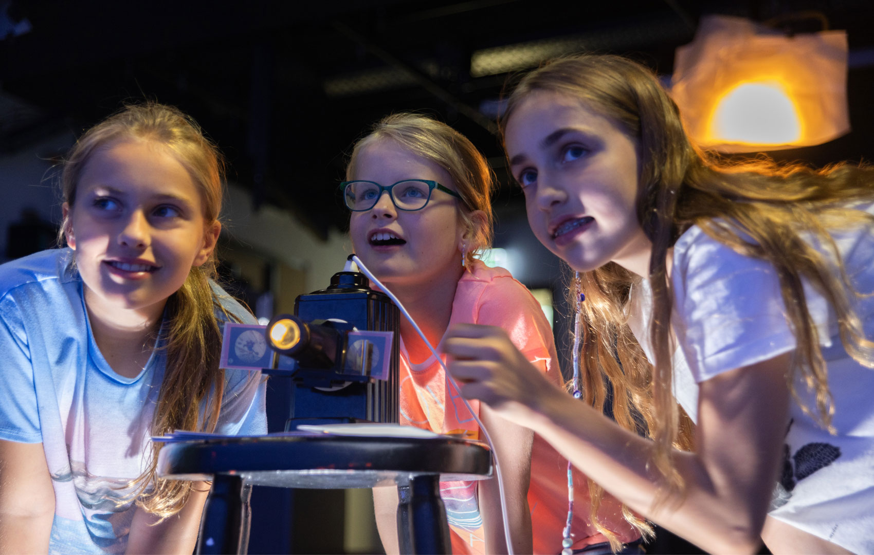 Drei Mädchen schauen aus dem Bild heraus. Vor ihnen steht ein kleiner Projektor, in dem ein mit Bildern bedruckter Streifen steckt. Das Projektorlicht strahlt.