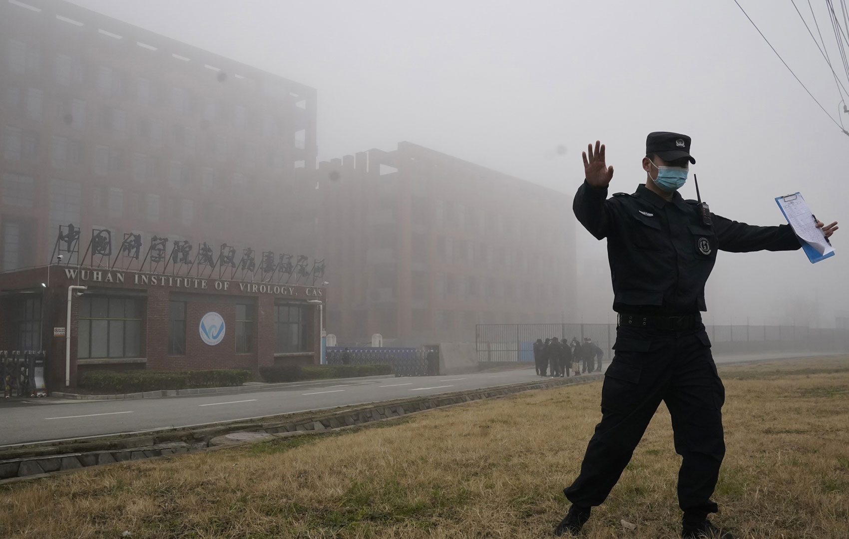 Ein Mann in Uniform und Maske läuft mit ausgestreckten Händen am rechten Bildrand in Richtung Unbekannter. In der einen Hand hält er ein Klemmbrett. Es scheint, als wollte er Menschen zurückhalten, näher zu kommen. Im Hintergrund steht ein Plattenbau mit der Aufschrift WUHAN INSTITUTE OF VIROLOGY.