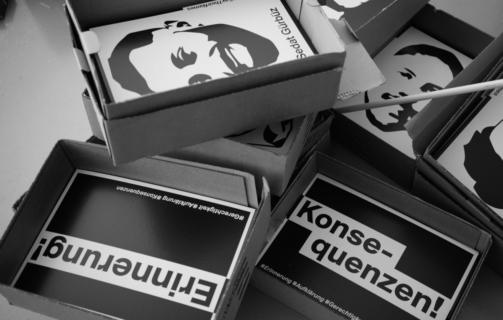 Auf einer Tischplatte liegen graue Pappkästen mit vergrößerten PersonenBildern. In zwei Kisten liegen auch Blätter, auf denen weiß auf schwarz folgende Wörter gedruckt sind: Erinnerung!, Konsequenzen!