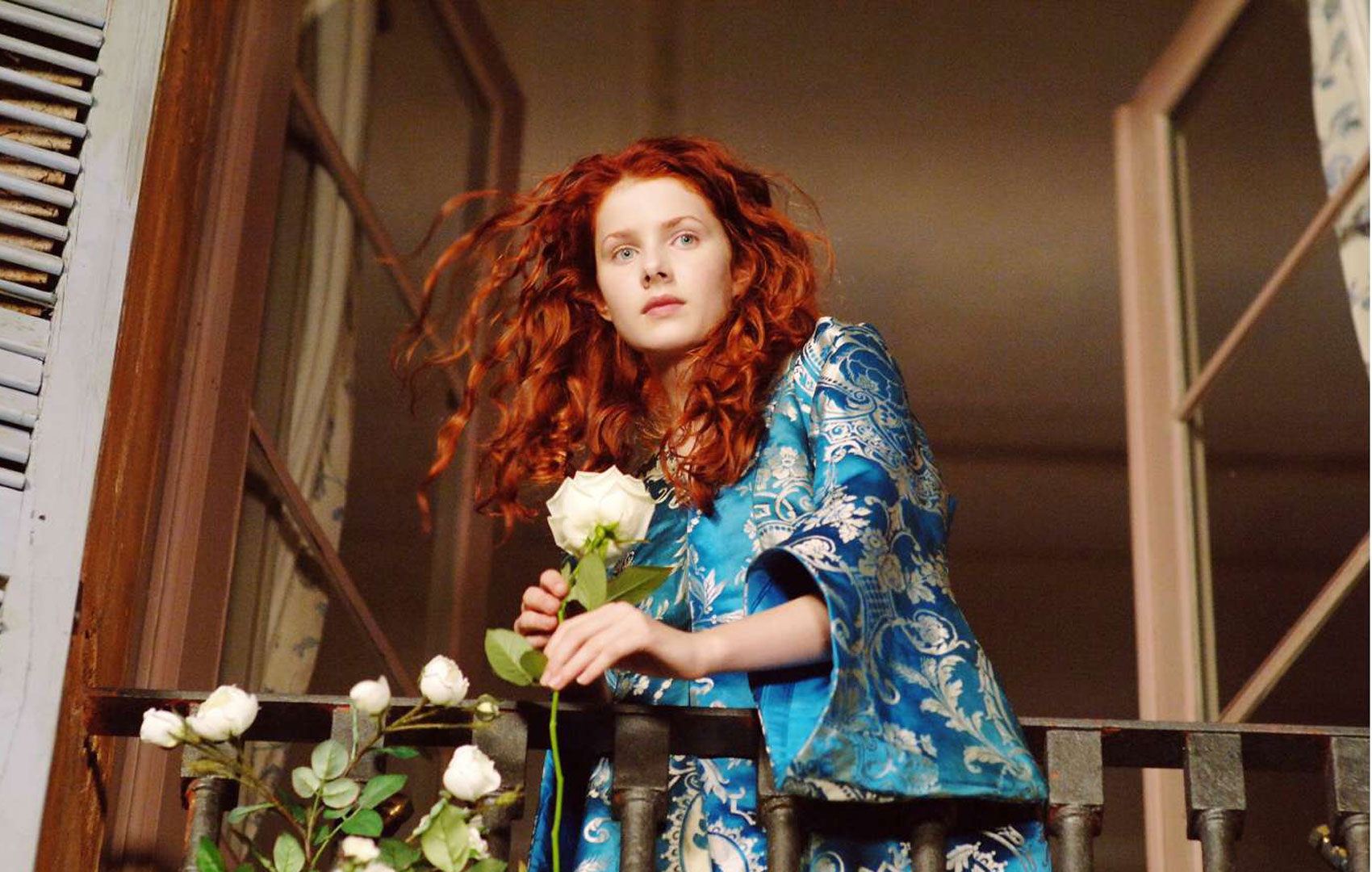 Eine junge Frau mit wallendem roten Haar und blauem Kleid schaut aus einem Balkonfenster nach unten. Sie ist etwas nach vorne gebäugt und hält eine langstielige, rote Rose in der Hand.