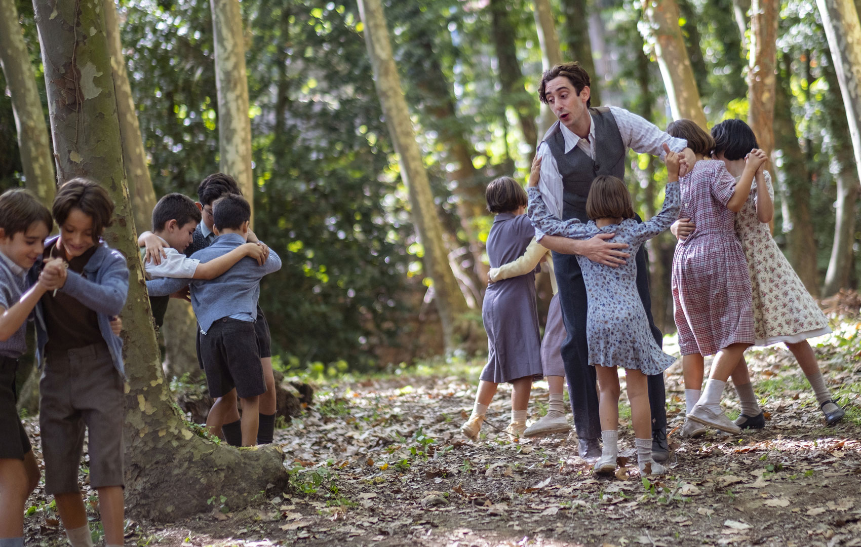 Kinder tanzen in einem Wald zwischen Baumstämmen miteinander. Sie tanzen immer in Paaren. Ein Kind tanzt mit einem erwachsenen Mann.