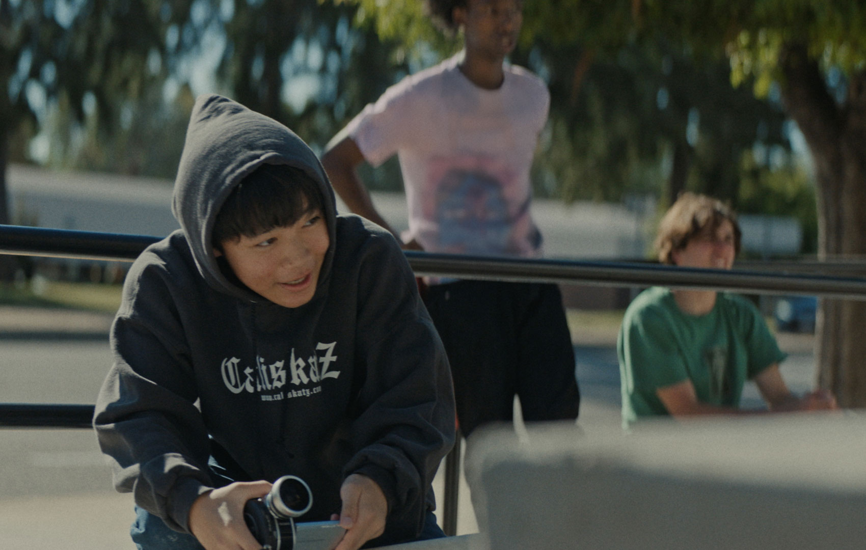 Ein Junge im Kapuzepulli sitzt in der Hocke am vorderen, linken Bildrand. Er schaut nach rechts und hält eine Kamera auf Bauchhöhe in der Hand. Im Hintergrund sind unscharf zwei weitere Jungs zu sehen.