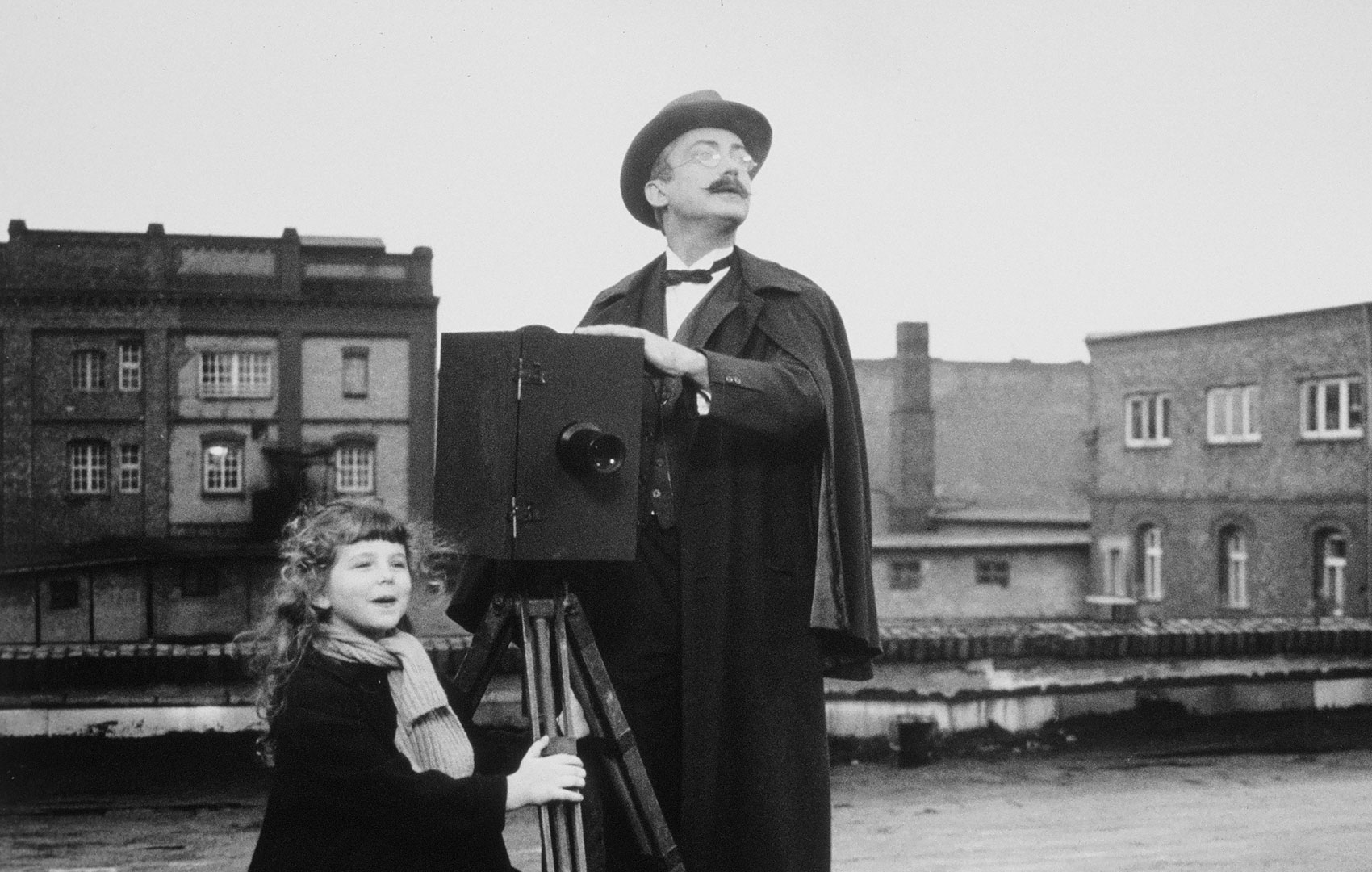 Ein Mann steht neben einer kastigen, altmodischen Kamera. Links neben ihm steht ein kleines Mädchen und hält sich am Stativ fest. Beide schauen rechts aus dem Bild hinaus. Im Hintergrund sieht man eine Häuserzeile. Das Bild ist schwarz-weiß.