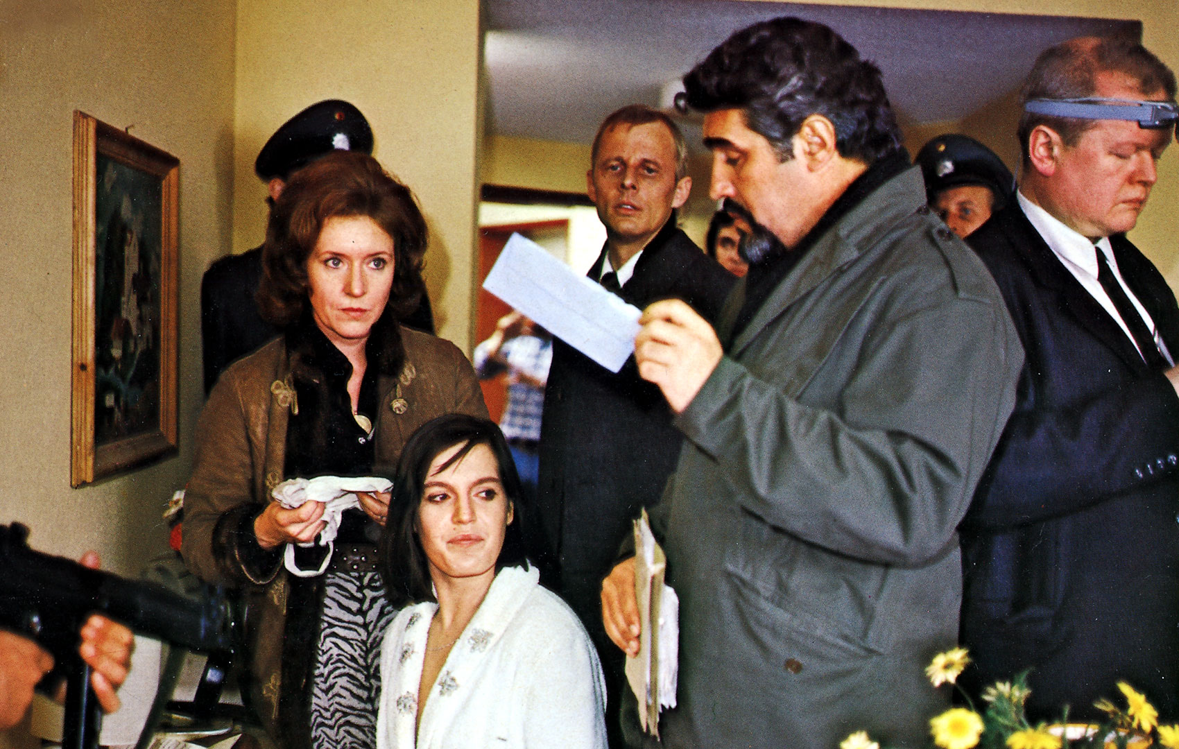 Eine Frau mit schwarzen Haaren und weißer Bluse ist umringt von Männern in Anzügen und einer weiteren Frau. Auf die Frau mit den schwarzen Haaren wird eine Waffe gehalten. Die Personen scheinen von der Polizei zu sein; einer hat eine charakteristische Mütze auf.