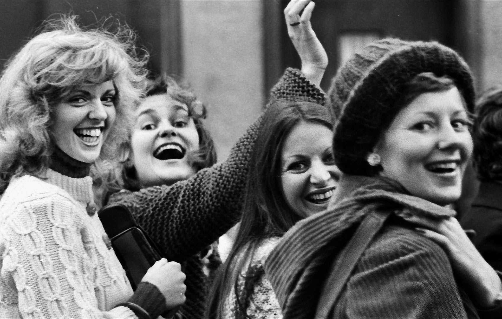 Schwarz-weiße Fotografie einer Seitenansicht von vier Frauen. Sie drehen ihre Köpfe zur Seite und lächeln in die Kamera.