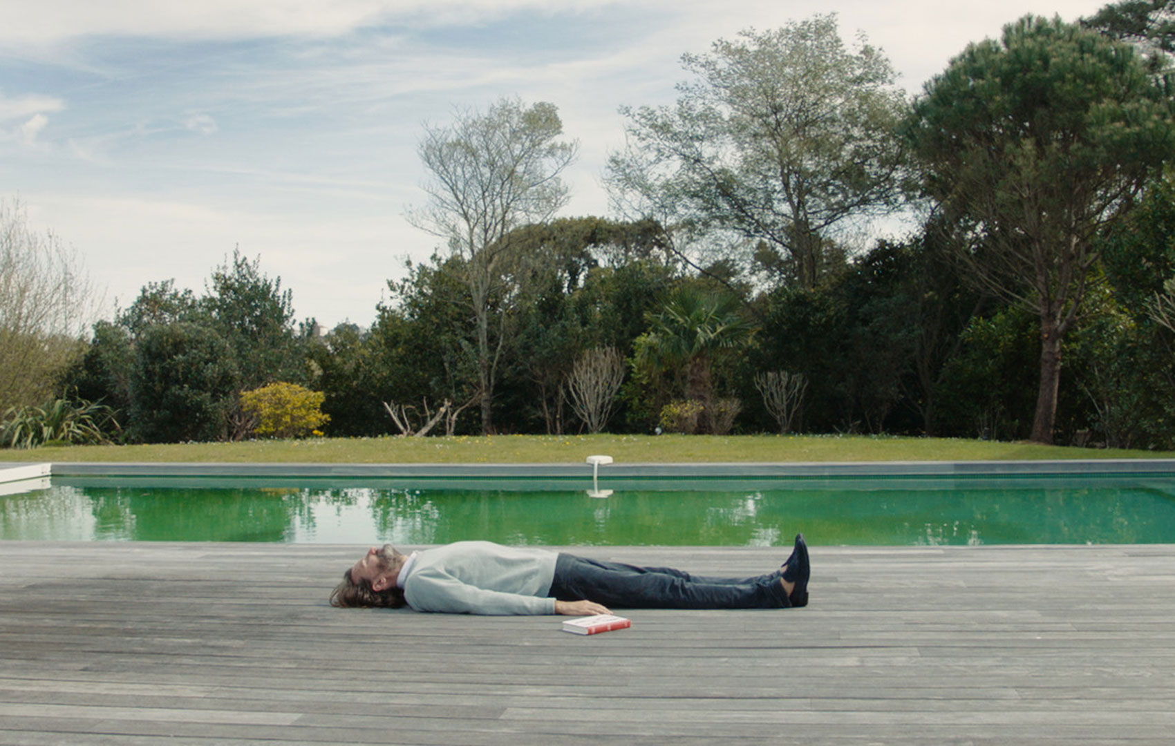 Ein Mann liegt komplett angezogen flach auf dem Holzboden vor einem großen Garten-Pool. Hinter dem Pool begrenzen eine Wiese und Bäume das Grundstück.