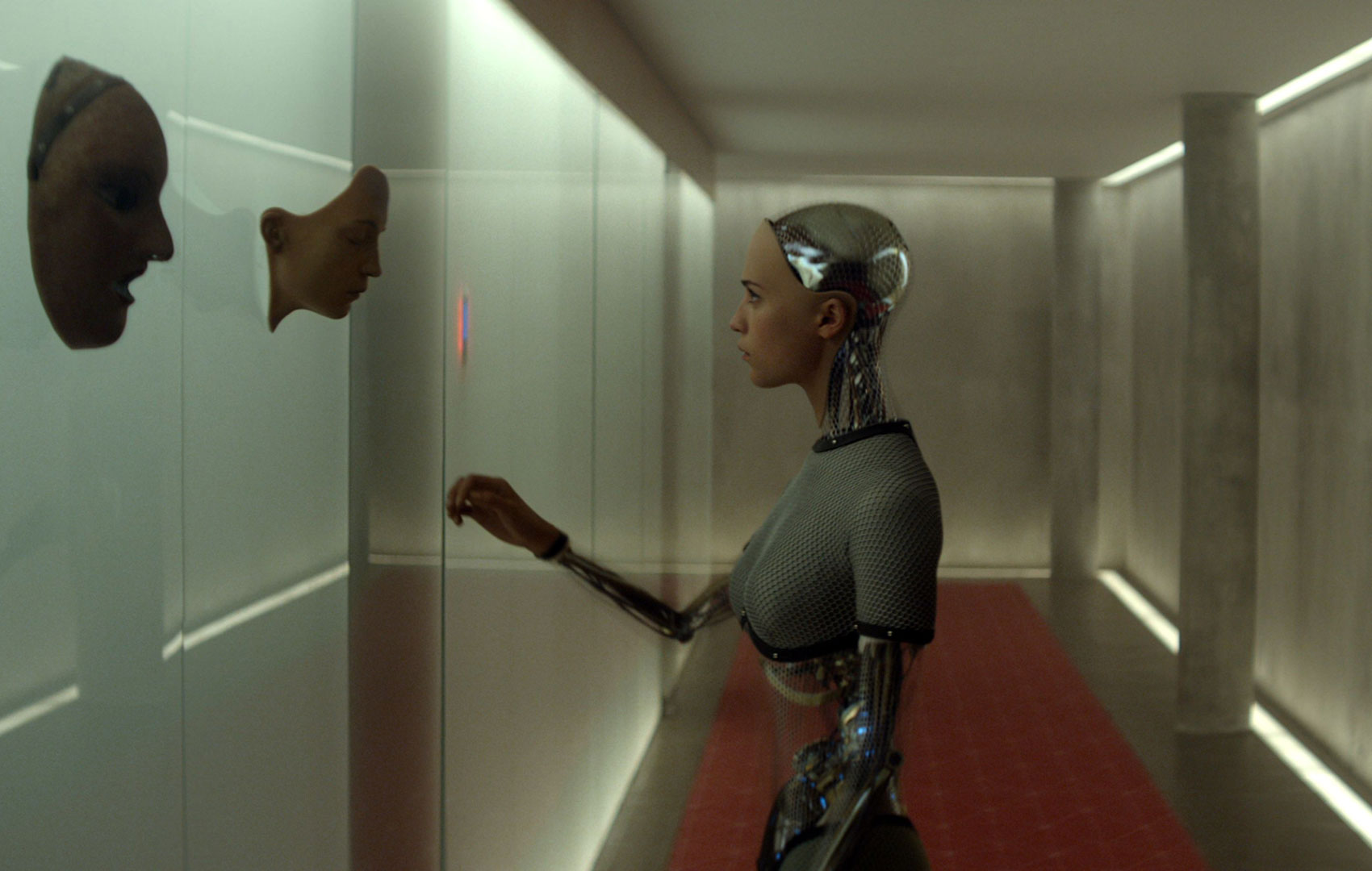 Eine Roboterfrau steht in einem Gang und schaut sich hautfarbene Gesichtsmasken an, die an der Wand hängen. Sie hat die Hand ausgestreckt, als wollte sie eine anfassen.