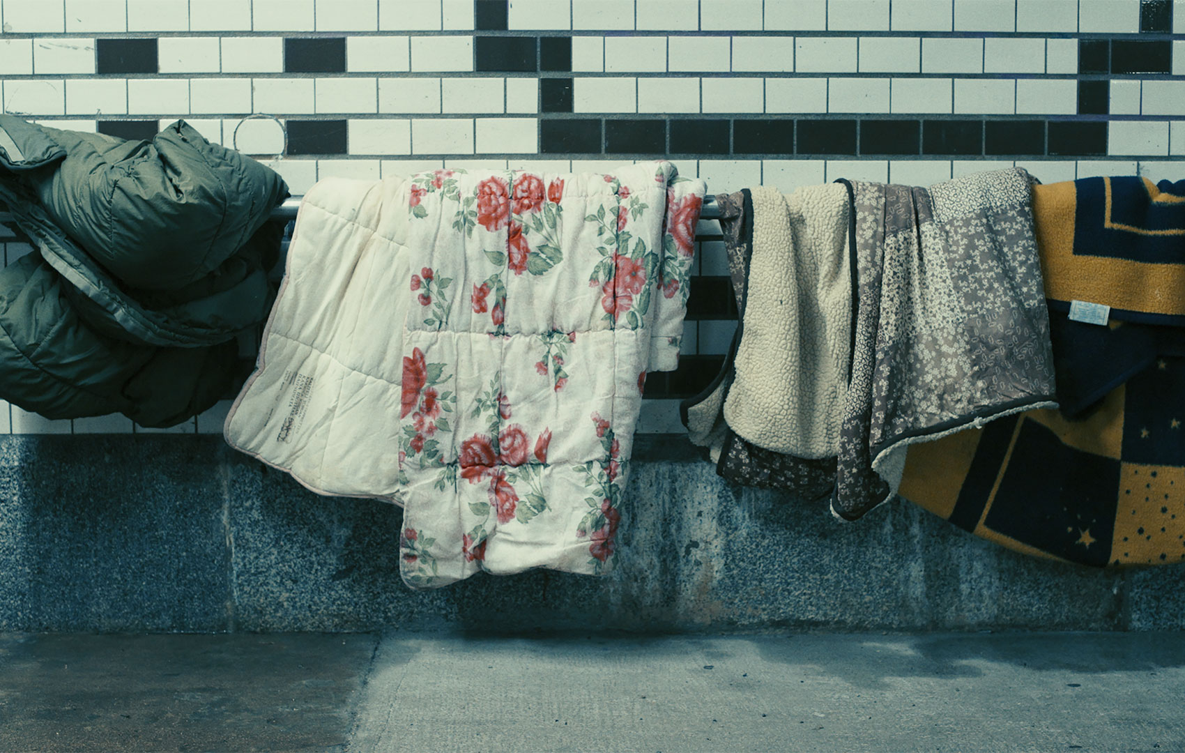 Unterschiedliche Decken hängen nebeneinander über einem Geländer vor einer Wand, die weiß gefliest ist und unten eine Steinabsetzung hat.