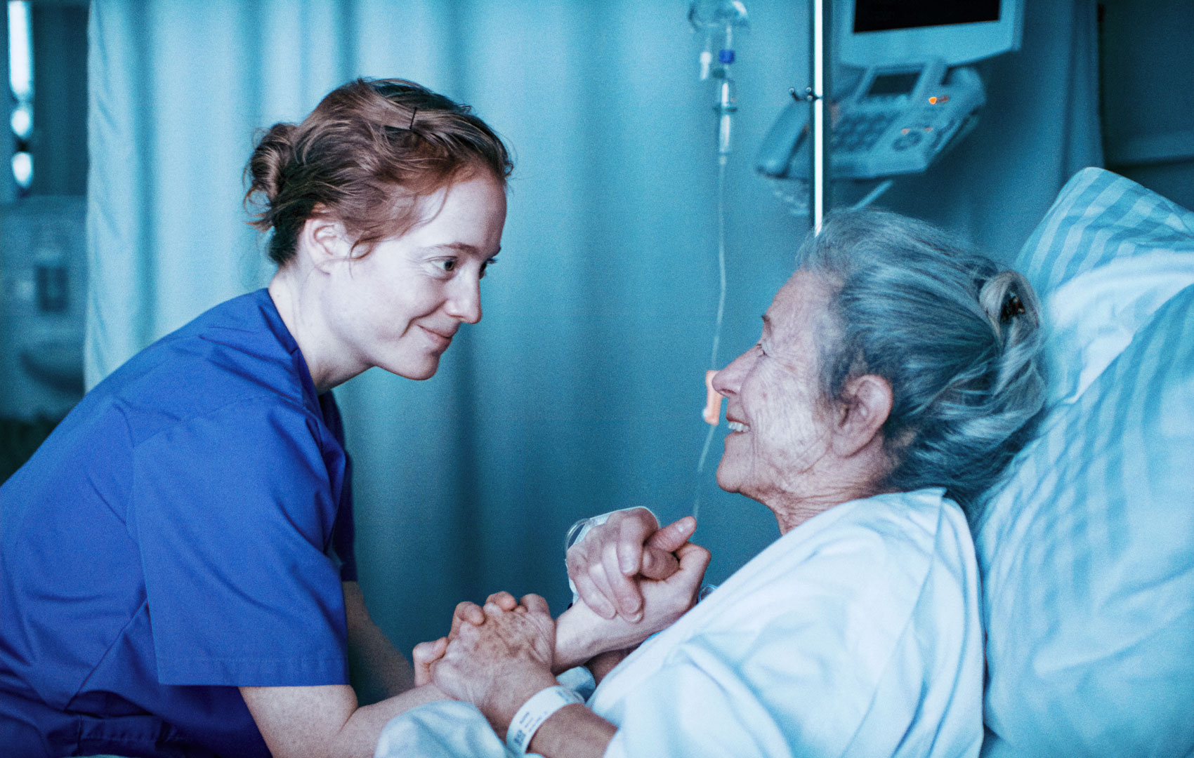Eine Krankenschwester sitzt am Bett einer alten Frau. Sie lächeln sich an und geben sich die Hände.