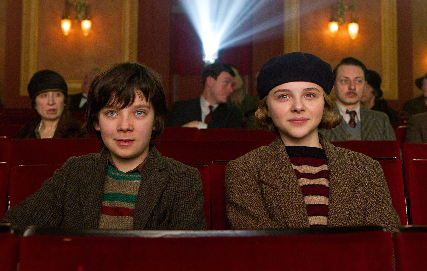 Ein Mädchen und ein Junge sitzen in einem Kinosaal auf roten Plüschsesseln. Im Hintergrund sieht man, wie das Licht aus dem Projektor in den Saal strahlt. Beide schauen freudig-erwartungsvoll.