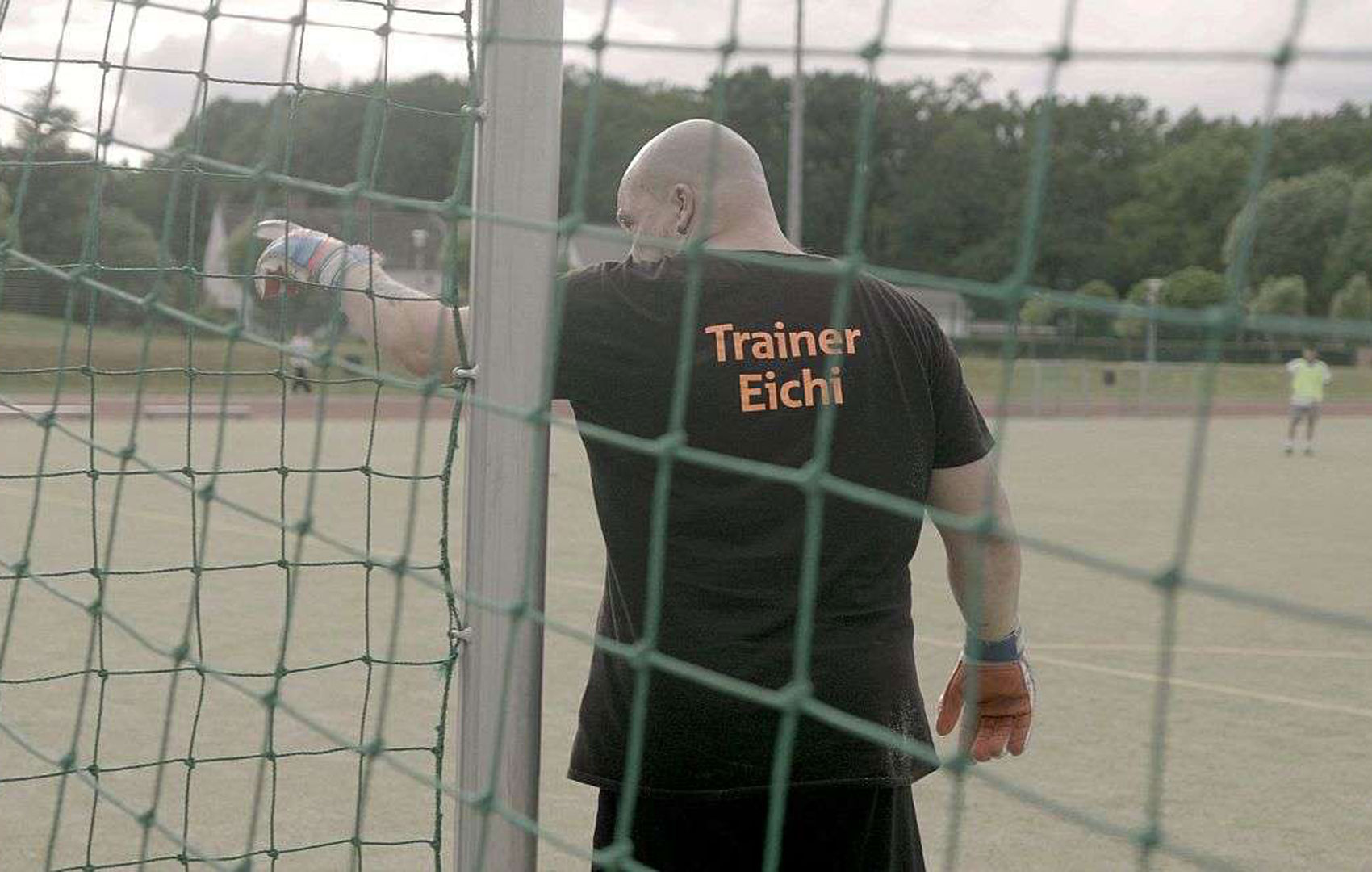 Durch ein Fußballtor-Netz hindurch sieht man den Rücken eines Mannes, der mit ausgestrecktem Arm nach links deutet. Auf seinem schwarzen T-Shirt steht: Trainer Eichi.
