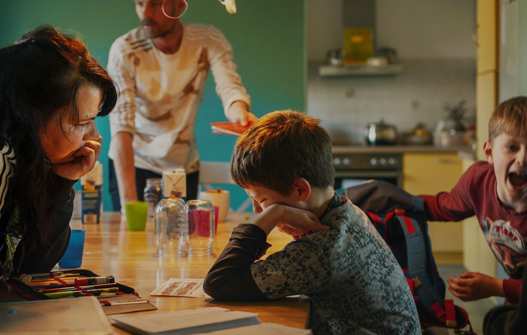 Ein Junge sitzt mit den Ellenbogen auf den Tisch gesützt. Man sieht ihn von der Seite, sein Gesicht ist durch die Hand fast verdeckt. Ihm gegenüber sitzt in gleicher Pose eine erwachsene Frau. Am rechten Bildrand hinter dem Jungen sieht man noch einen zweiten, der schreit. Am Tischend steht ein weiterer Erwachsener.