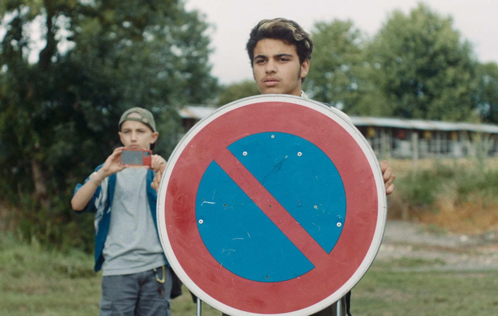 Ein Junge hat ein großes Verkehrschild in der Hand. Es ist das Zeichen für eingeschränktes Halteverbot. Etwas hinter ihm steht ein weiterer Junge und filmt mit dem Handy.