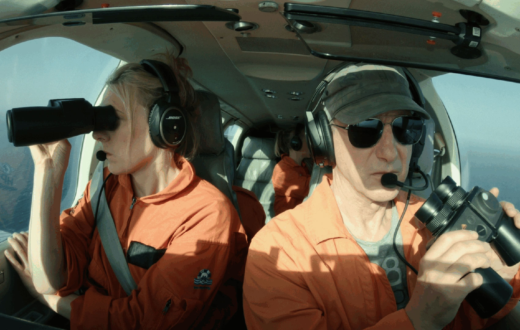 Zwei Erwachsene in orangefarbenen Overalls sitzen im Hubschrauber und schauen mit Ferngläsern nach draußen.