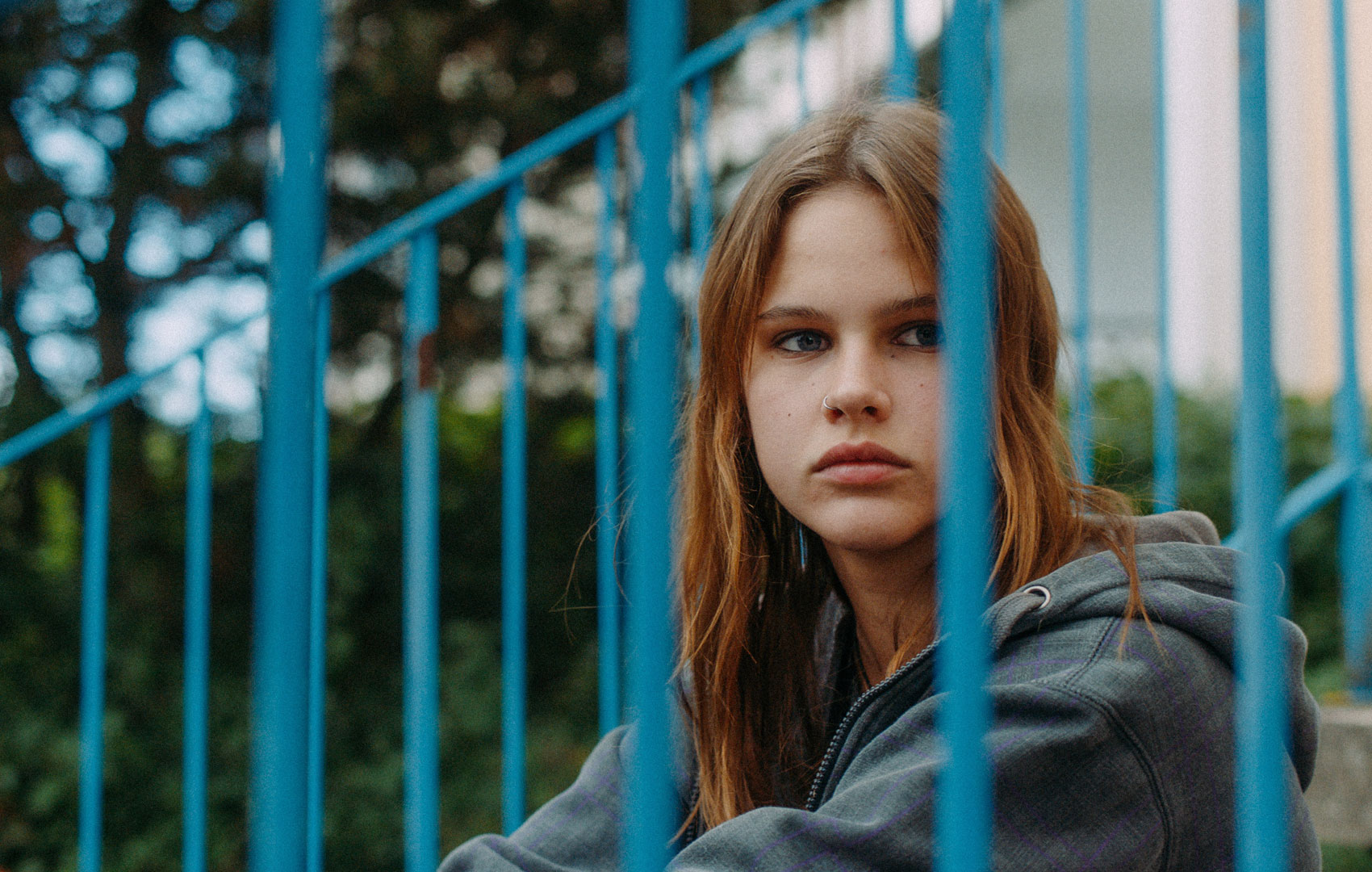 Ein Mädchen sitzt auf einer Treppe hinter einem blauen Treppengeländer. Man sieht ihren Kopf eingerahmt von der Gitterstäben. Sie schaut aus dem Bild hinaus und sieht unglücklich aus.